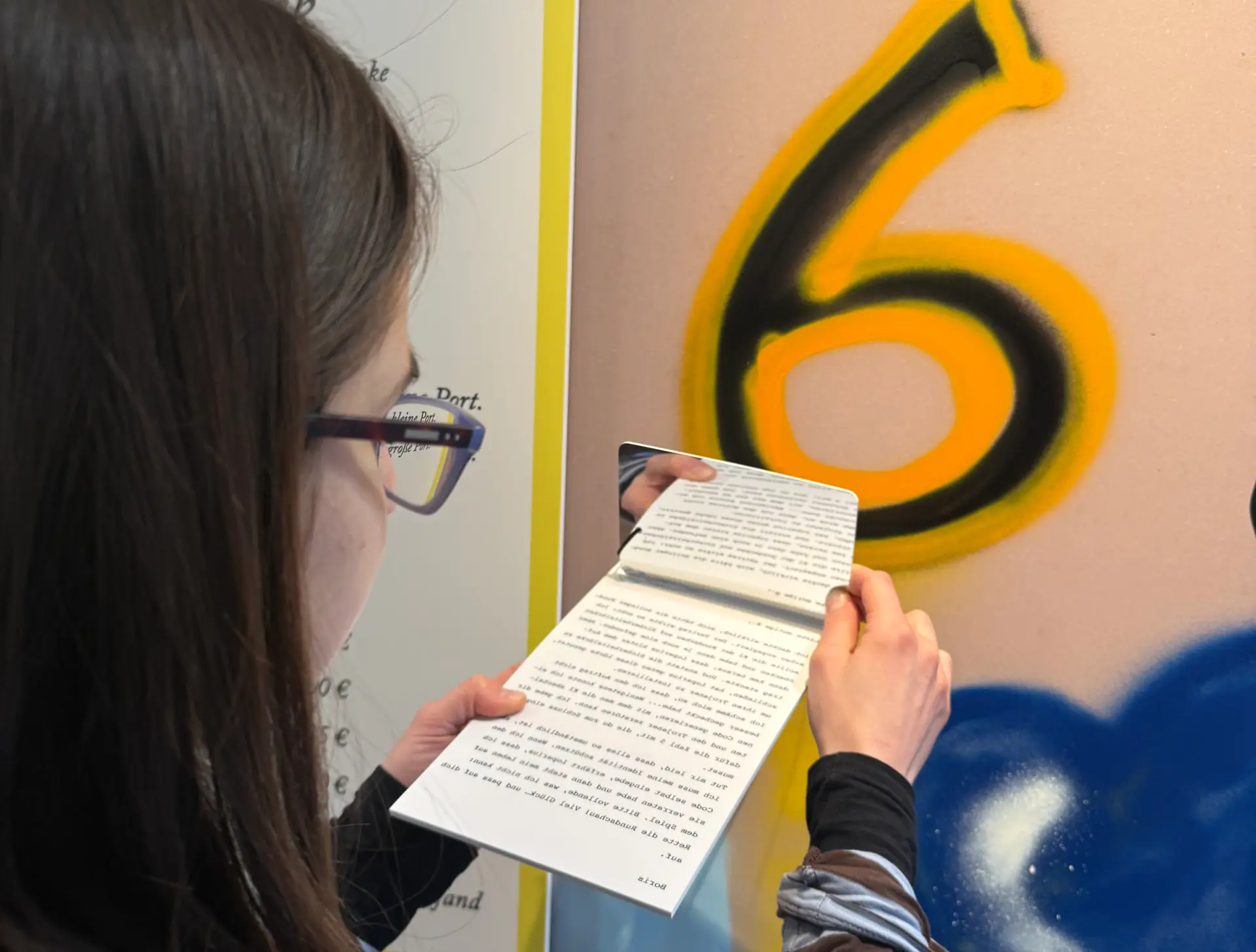 Eine Frau mit Brille und schulterlangen Haaren hat einen Text in der Hand, der verkehrt geschrieben ist. Mit einem Spiegel liest sie den Text.