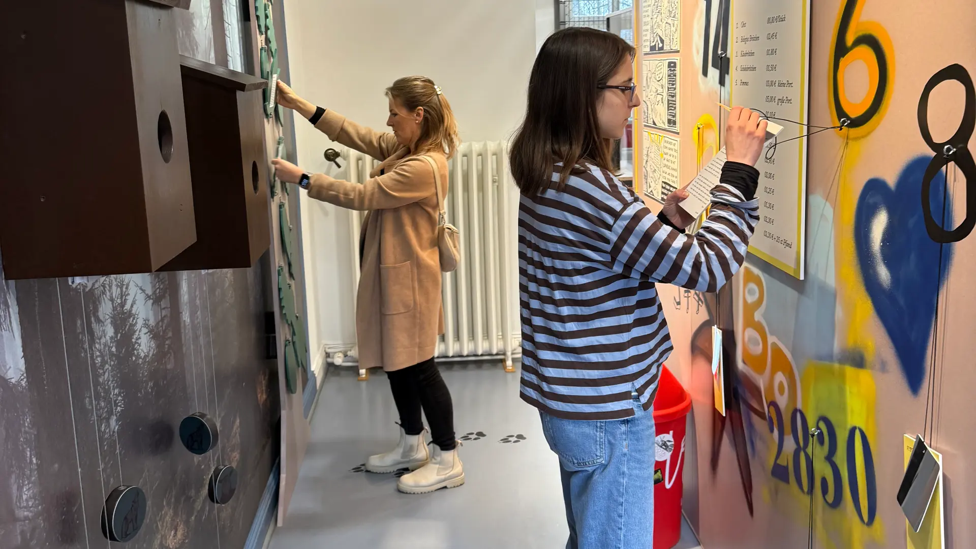 Zwei Frauen stehen in einem Ausstellungsraum. Eine Frau sieht sich einen Hut, der an der Decke hängt, an. Die zweite Frau liest einen Ausstellungstext.