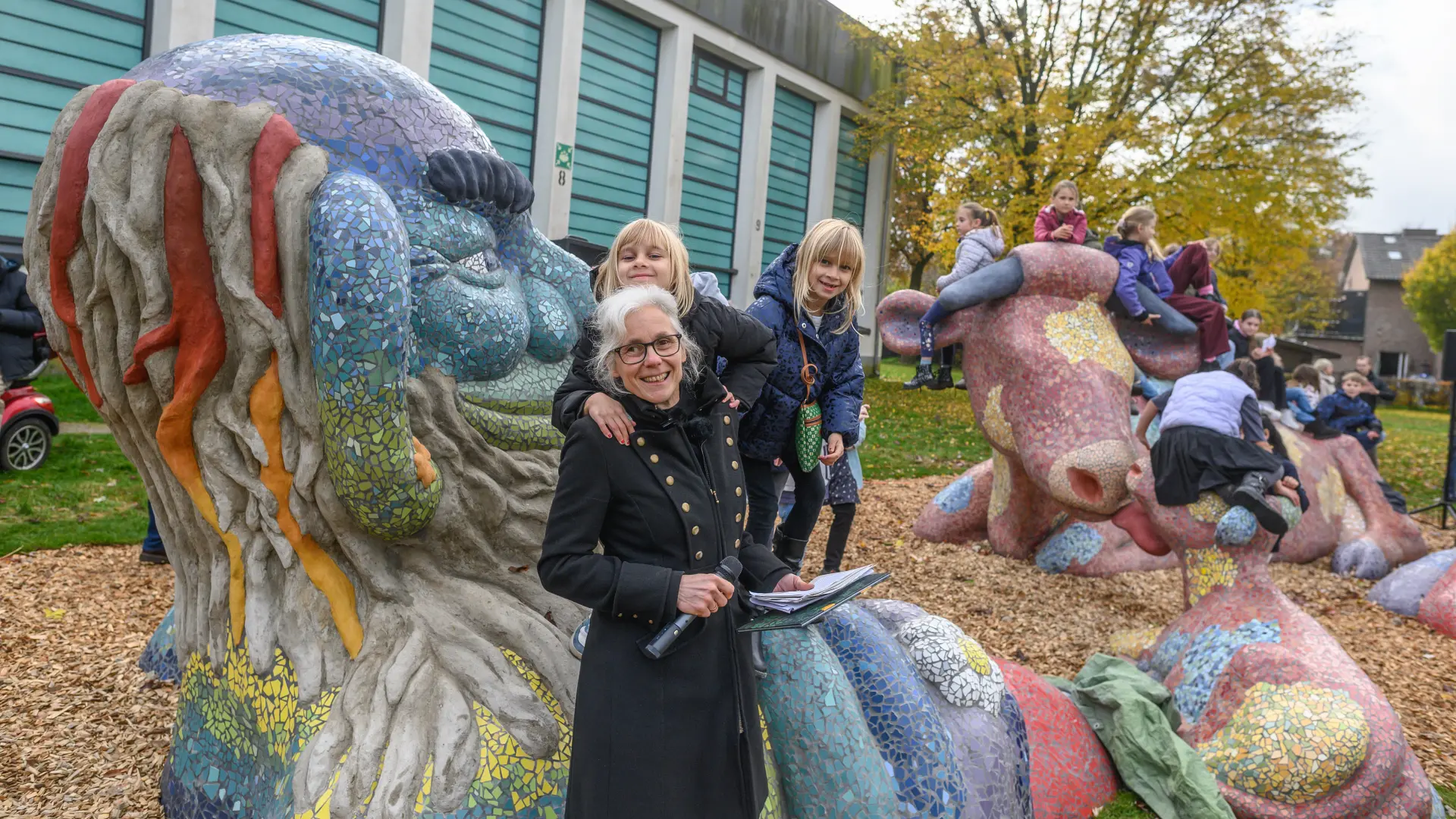 Eine Frau mit Brille, weißen Haaren und einem langen schwarzen Mantel hält ein Mikrofon in der Hand. Sie steht vor eine bunten Großskulptur gemeinsam mit mehreren Kindern, die auf der Skulptur spielen.
