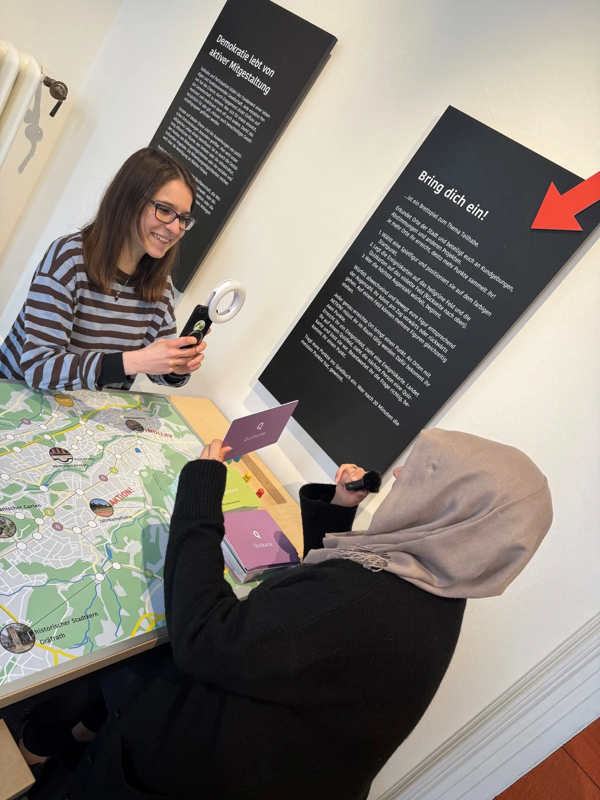 Eine Frau mit Brille und schulterlangen Haaren und eine frau mit Kopftuch sitzen an einem Spieltisch in einer Ausstellung. Sie drehen ein Interview mit Handy, Handylicht und einem Mini-Mikrofon.