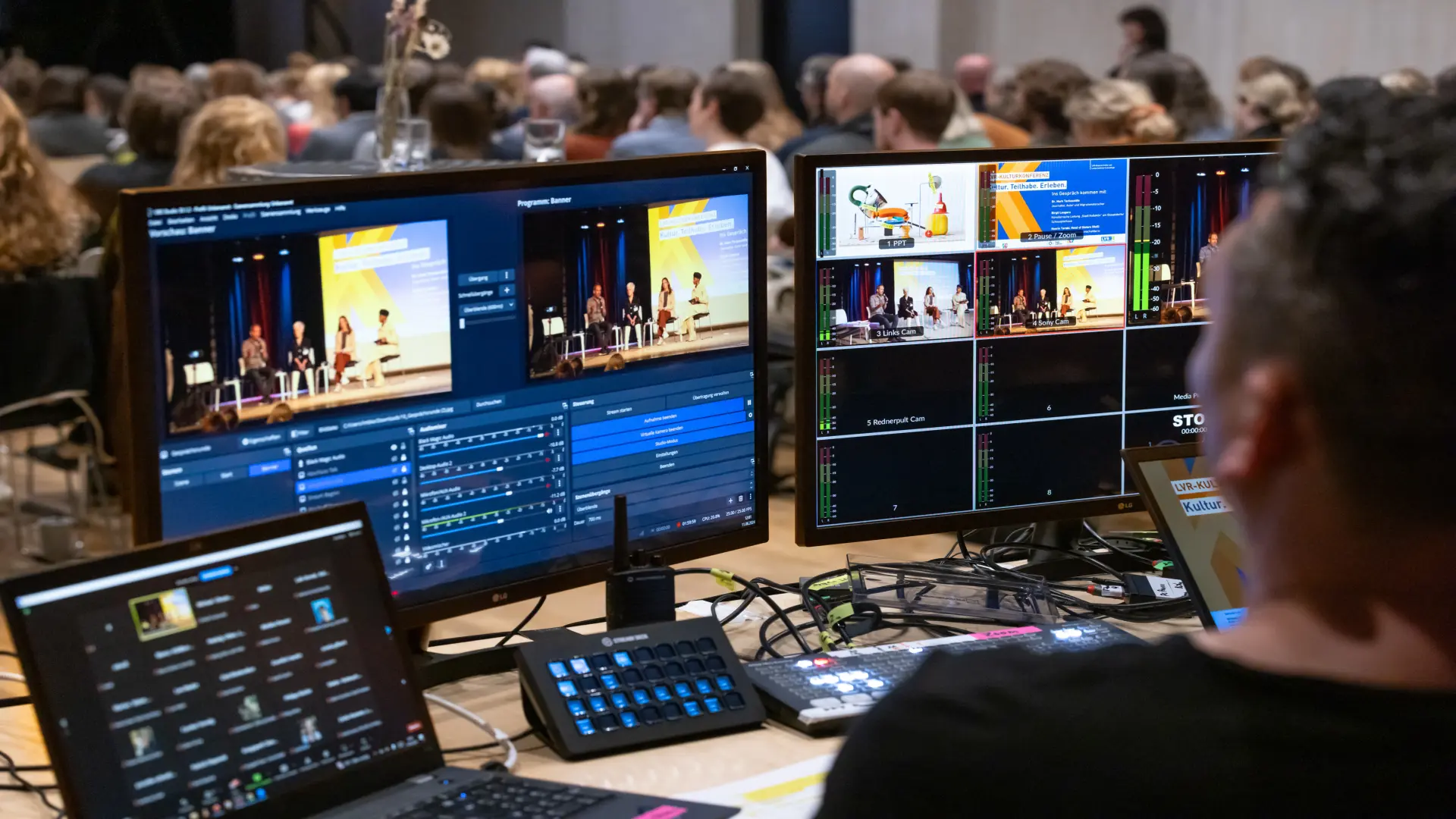 Foto: Blick über Technikpult mit mehreren Bildschirmen auf Publikum in einem großen Saal bei einer Präsentation.