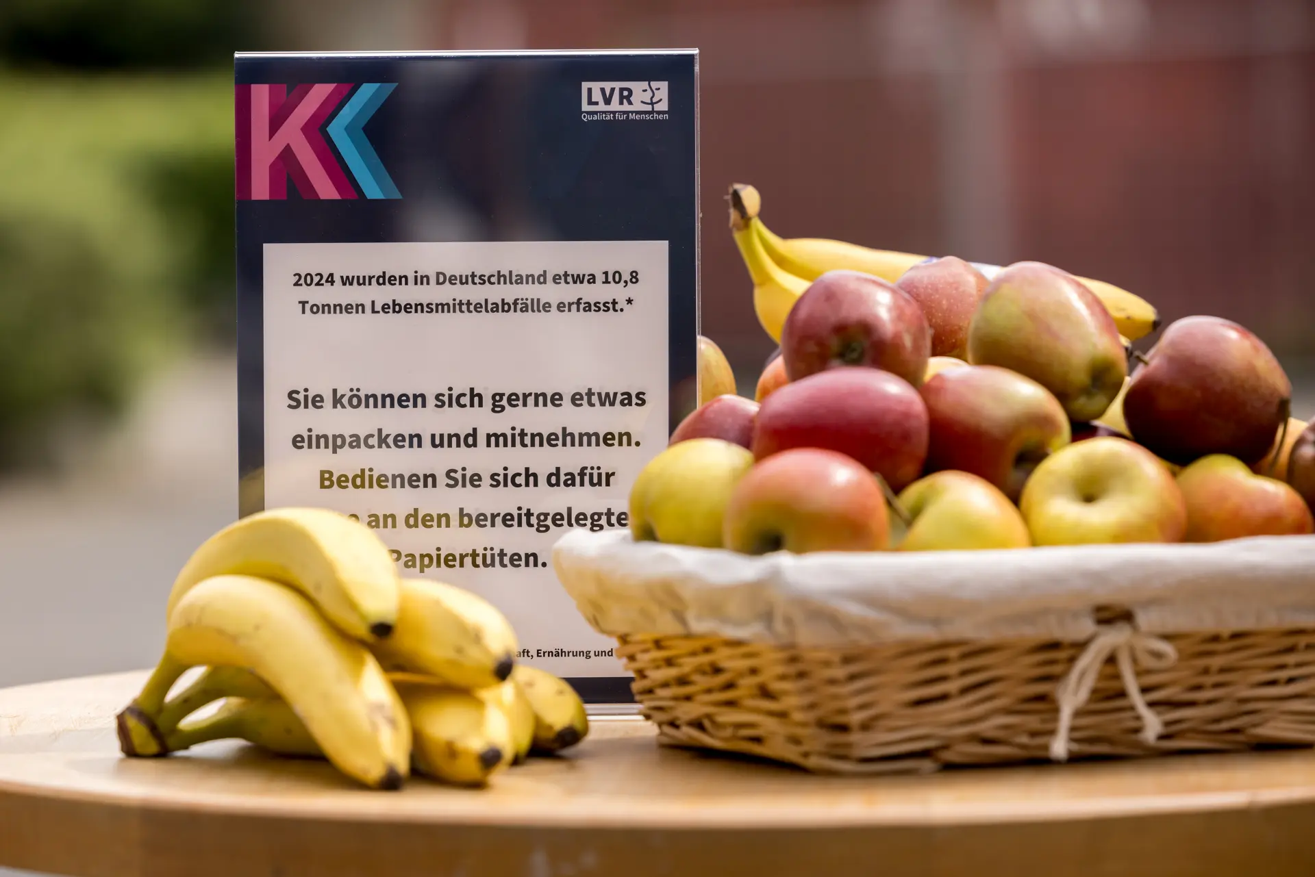 Foto: Korb mit Äpfeln und Birnen sowie Bananen auf einem Tisch neben einem Schild mit Text zur Lebensmittelverschwendung in Deutschland.
