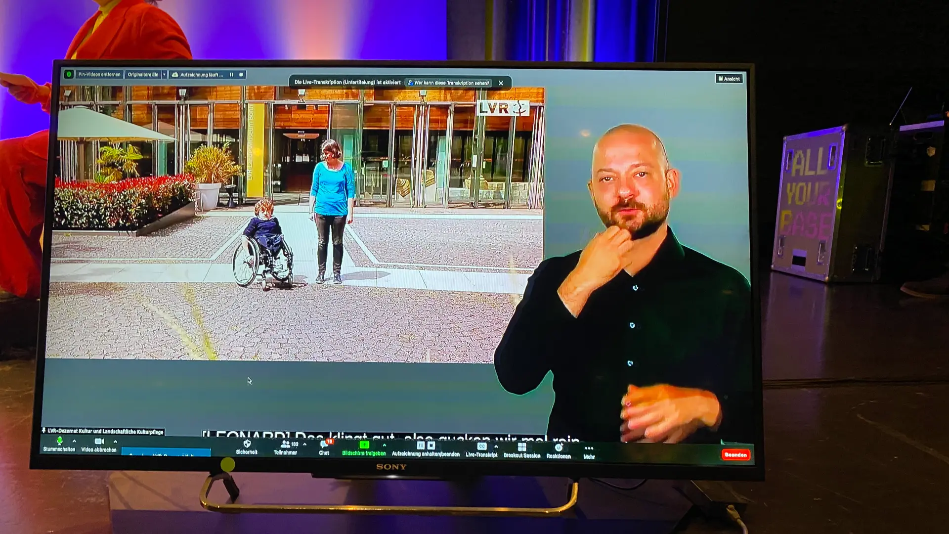 Foto: Fernsehbildschirm zeigt eine Szene mit einer Frau, die einen Rollstuhl schiebt, und einem Mann, der Gebärdensprache vor einem Gebäude mit Glasfassade verwendet.
