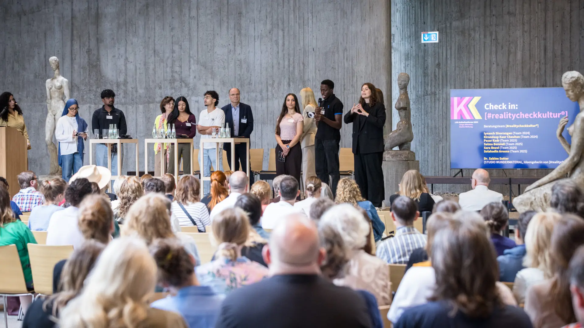 Foto: Mehrere Personen stehen auf einer Bühne vor einem Publikum in einem großen Raum mit Skulpturen und einem Bildschirm.
