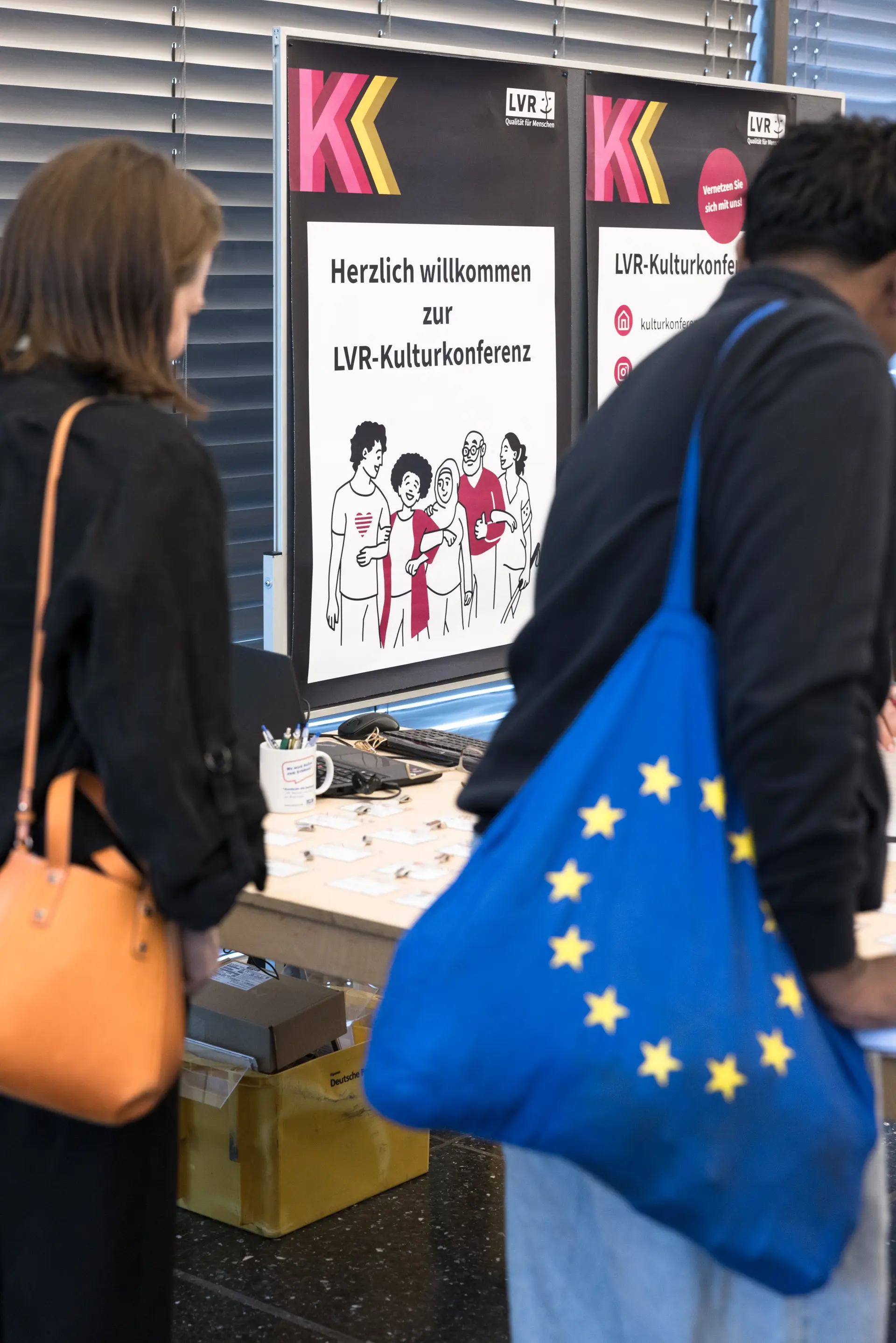 Foto: Zwei Personen mit Taschen stehen vor einem Tisch mit Informationsplakaten zur LVR-Kulturkonferenz.