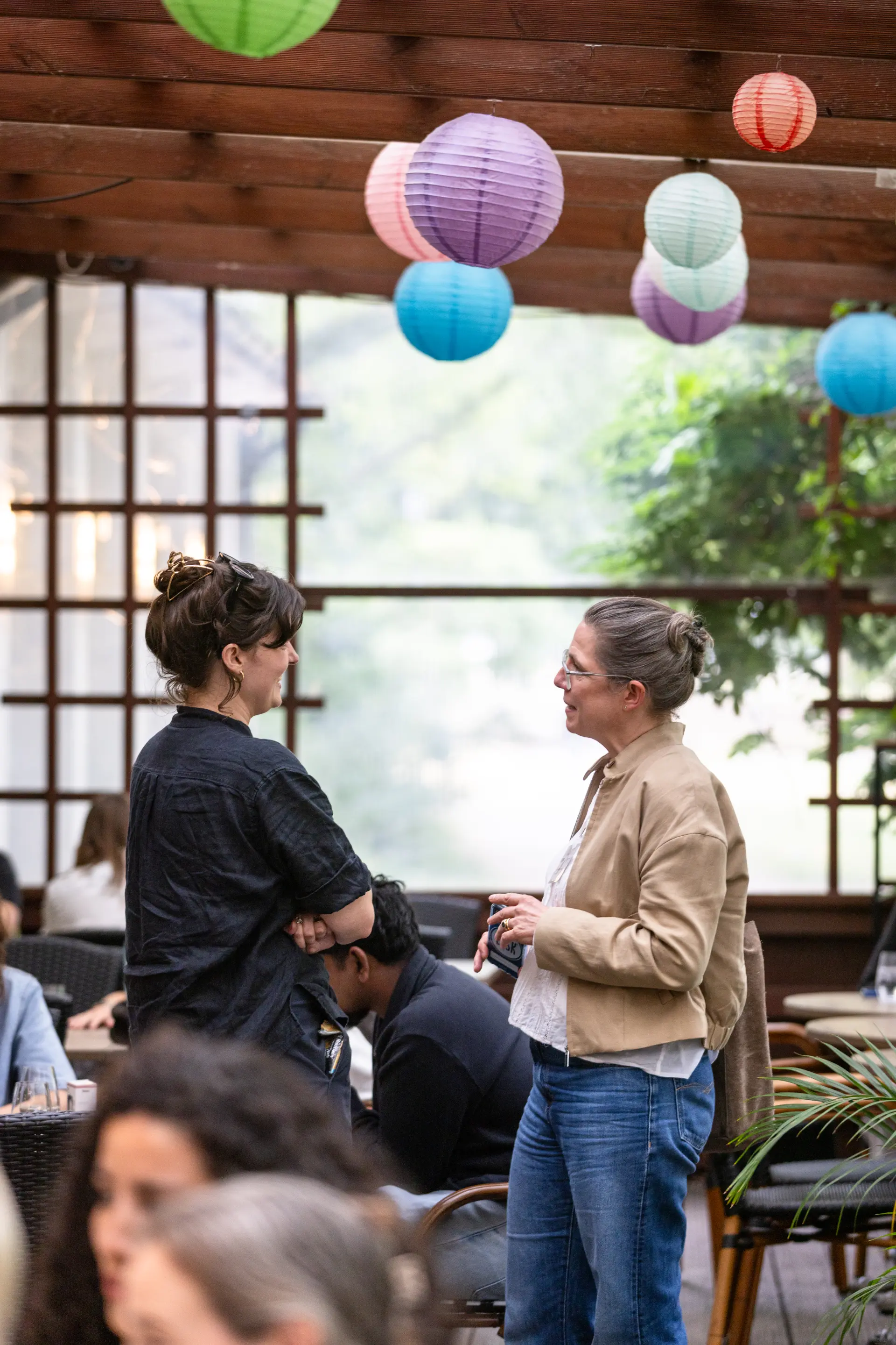 Foto: Zwei Frauen unter bunten Papierlaternen im Gespräch in einem Café mit Holzbalkendecke.