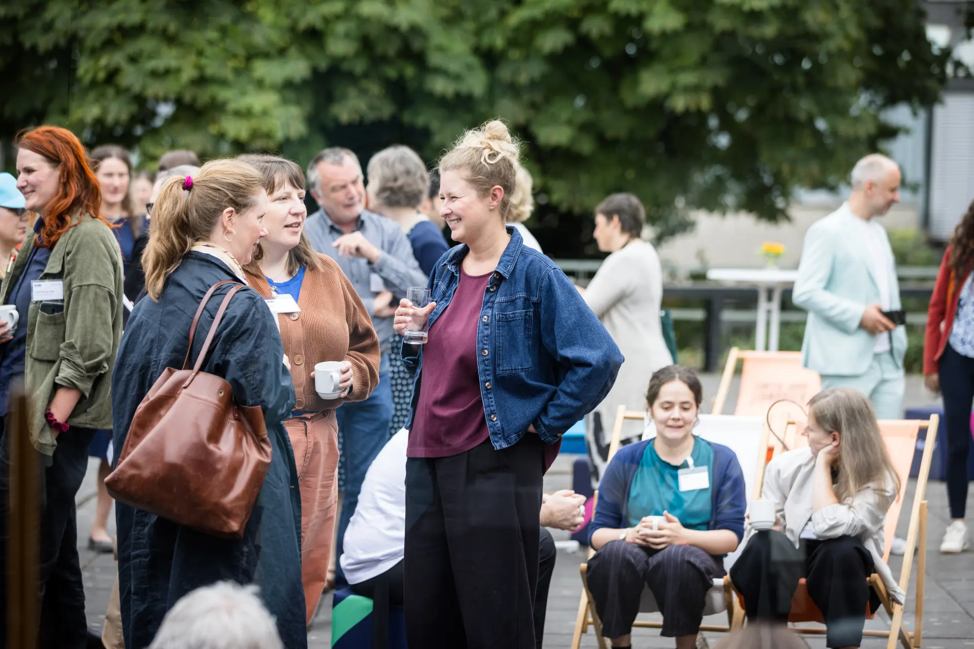 Foto: Gruppe von Menschen steht und sitzt im Freien, unterhält sich und hält Getränke in der Hand.