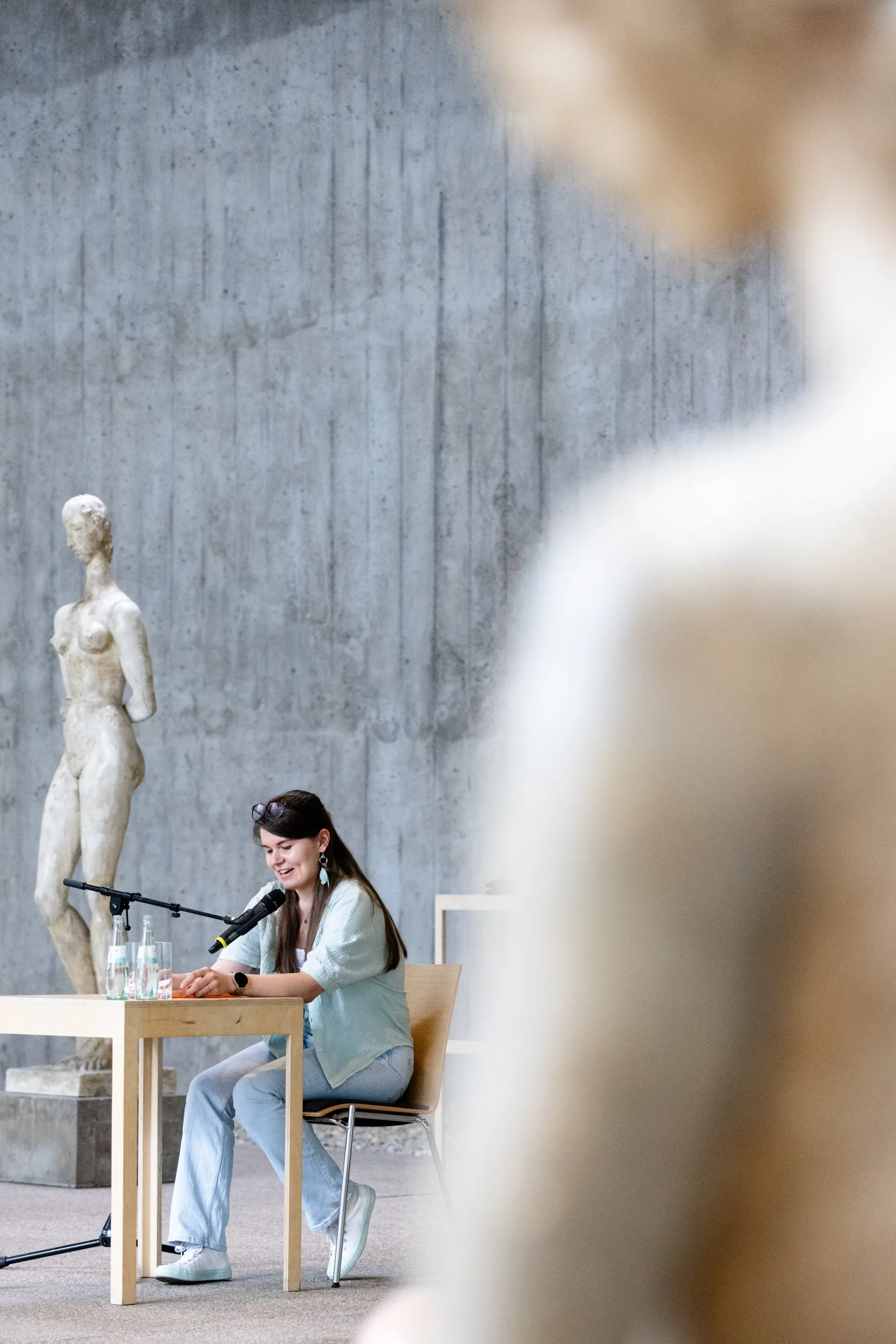Foto: Frau sitzt an einem Holztisch mit Mikrofon vor einer großen weißen Skulptur in einem Raum mit Betonwand.