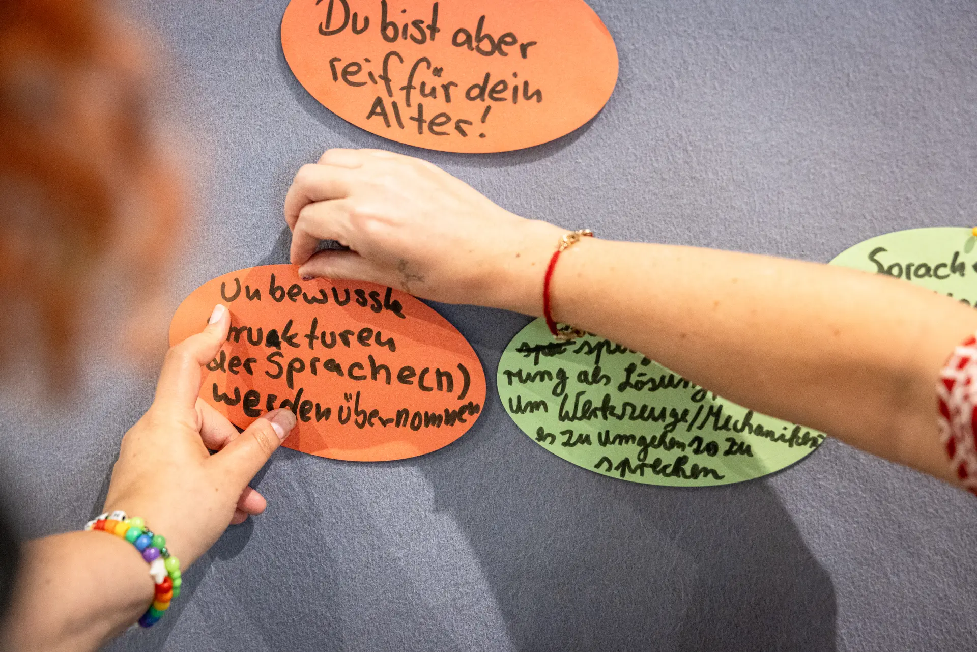 Foto: Zwei Hände heften bunte, ovale Zettel mit handgeschriebenem Text an eine Wand.