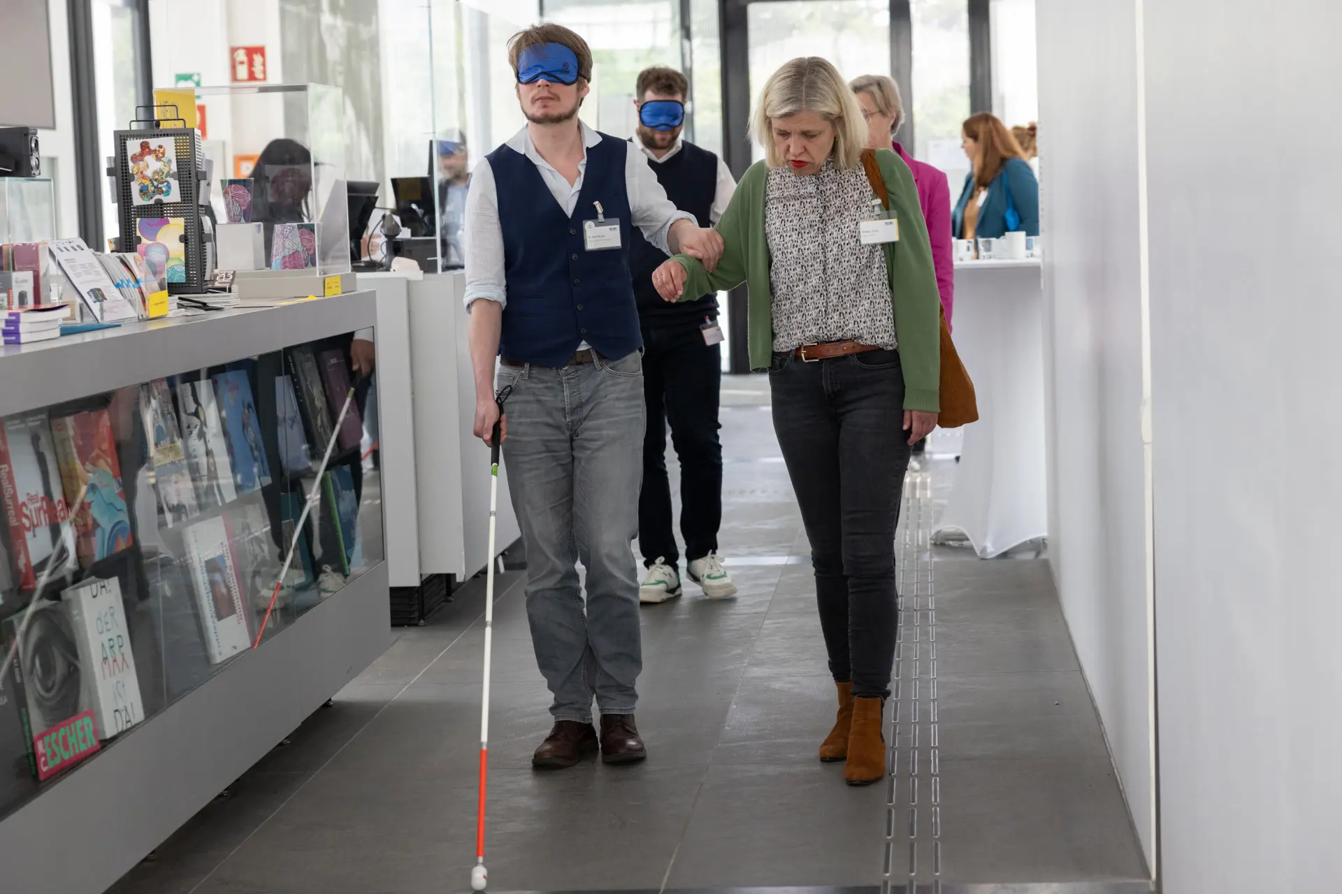 Foto: Zwei Personen gehen in einem hellen Flur, eine davon mit Blindenstock, die andere hält ihre Hand zur Unterstützung.