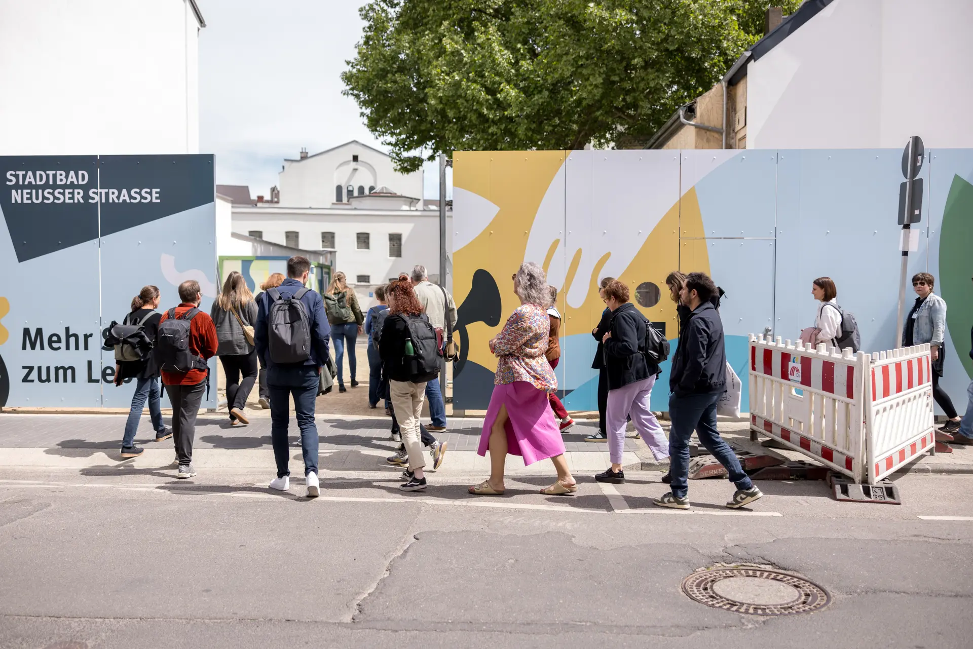 Foto: Gruppe von Menschen überquert eine Straße vor einer Baustellenabsperrung mit farbiger Wand und Schriftzug 'STADTBAD NEUSSER STRASSE"