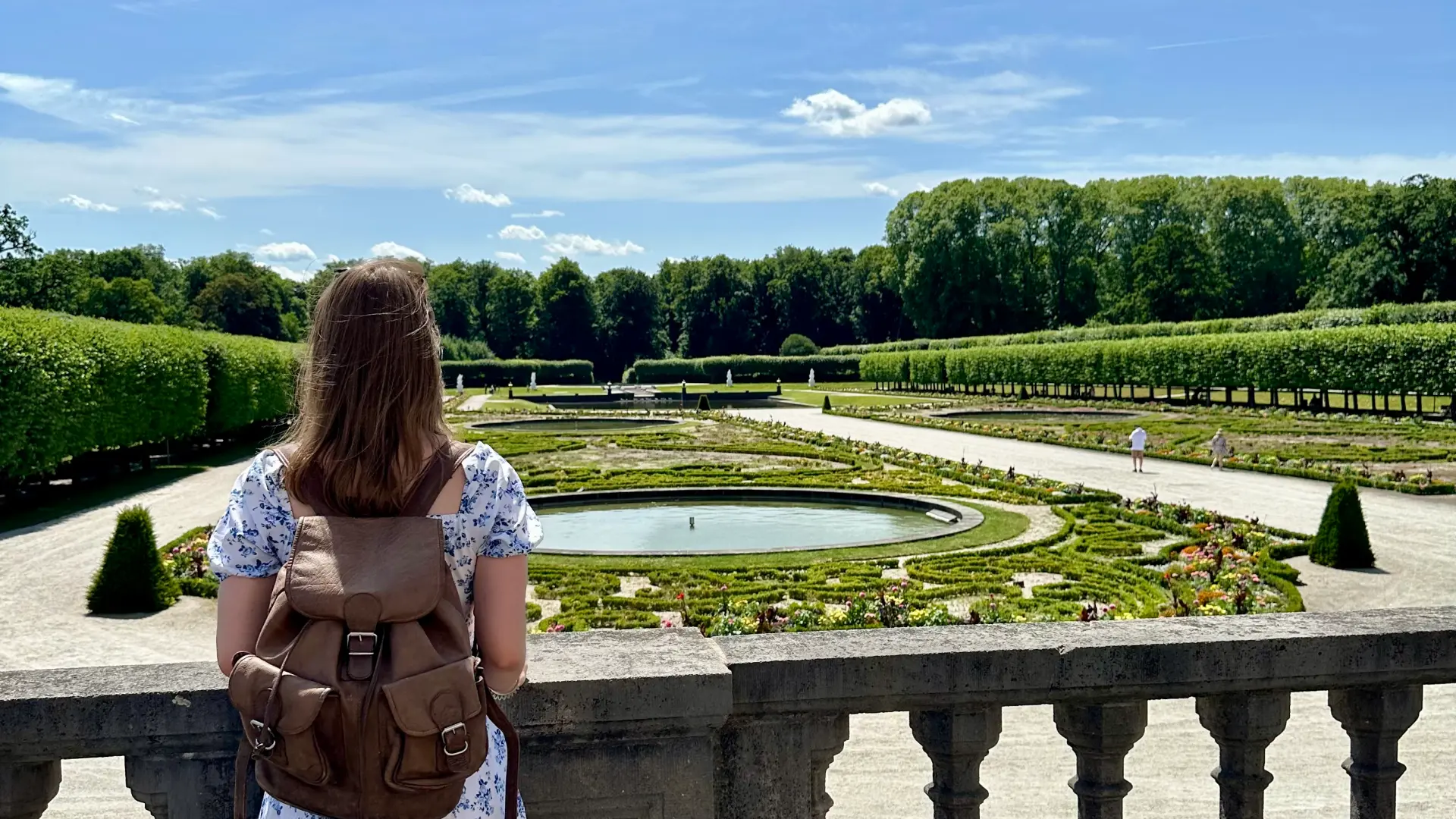 Foto: Eine Person steht an einem Balkongeländer und blick auf einen Schlossgarten mit Blumenbeeten, die Ornament ähnlich angepflanzt sind und in dessen Mitte ein Brunnen ist. Wege führen durch den Garten.