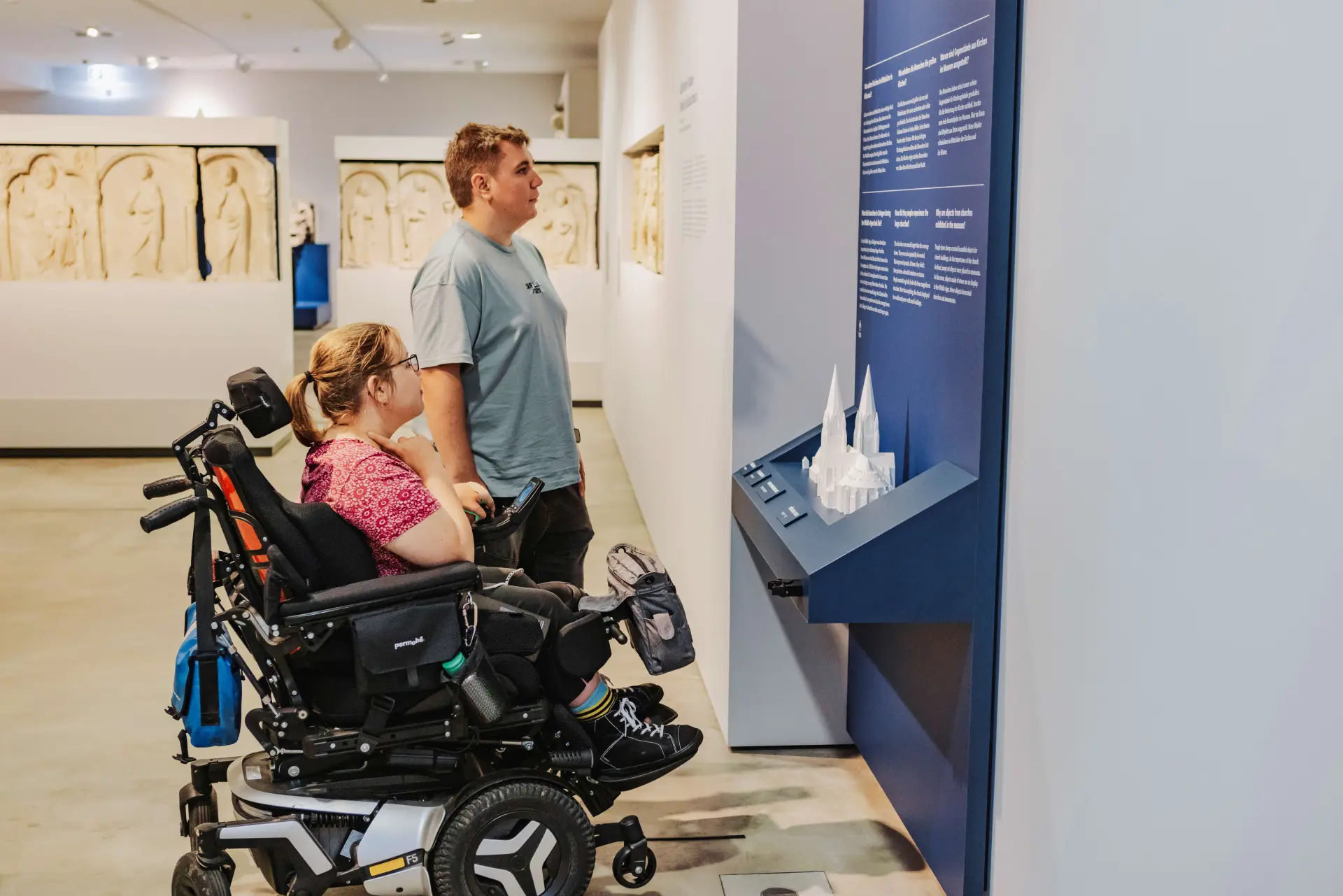 Foto: zwei Menschen betrachten ein Objekt in einem Museum. Eine Person davon sitzt in einem Elektro-Rollstuhl.