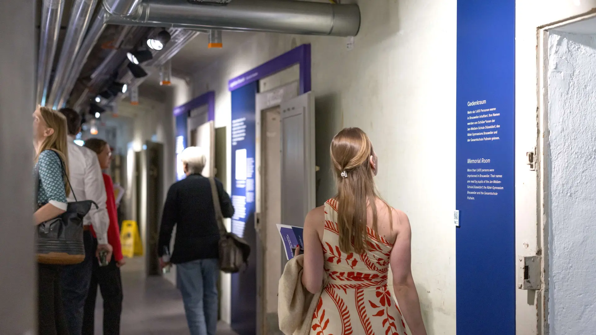 Foto: Mehrere Personen stehen in einem beleuchteten Kellergang und schauen sich eine Ausstellung an.