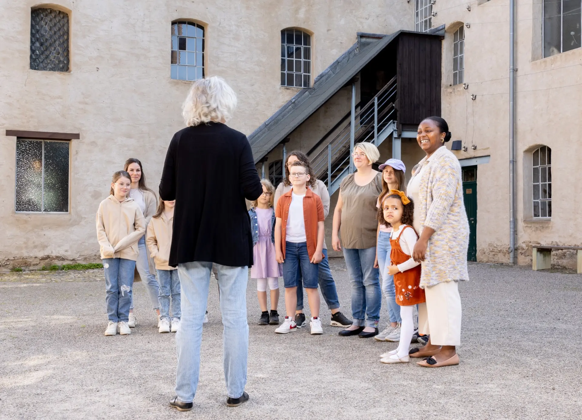 Foto: Eine Gruppe von Erwachsenen und Kindern steht vor einem Museum und hört einer Museumsmitarbeiterin zu.