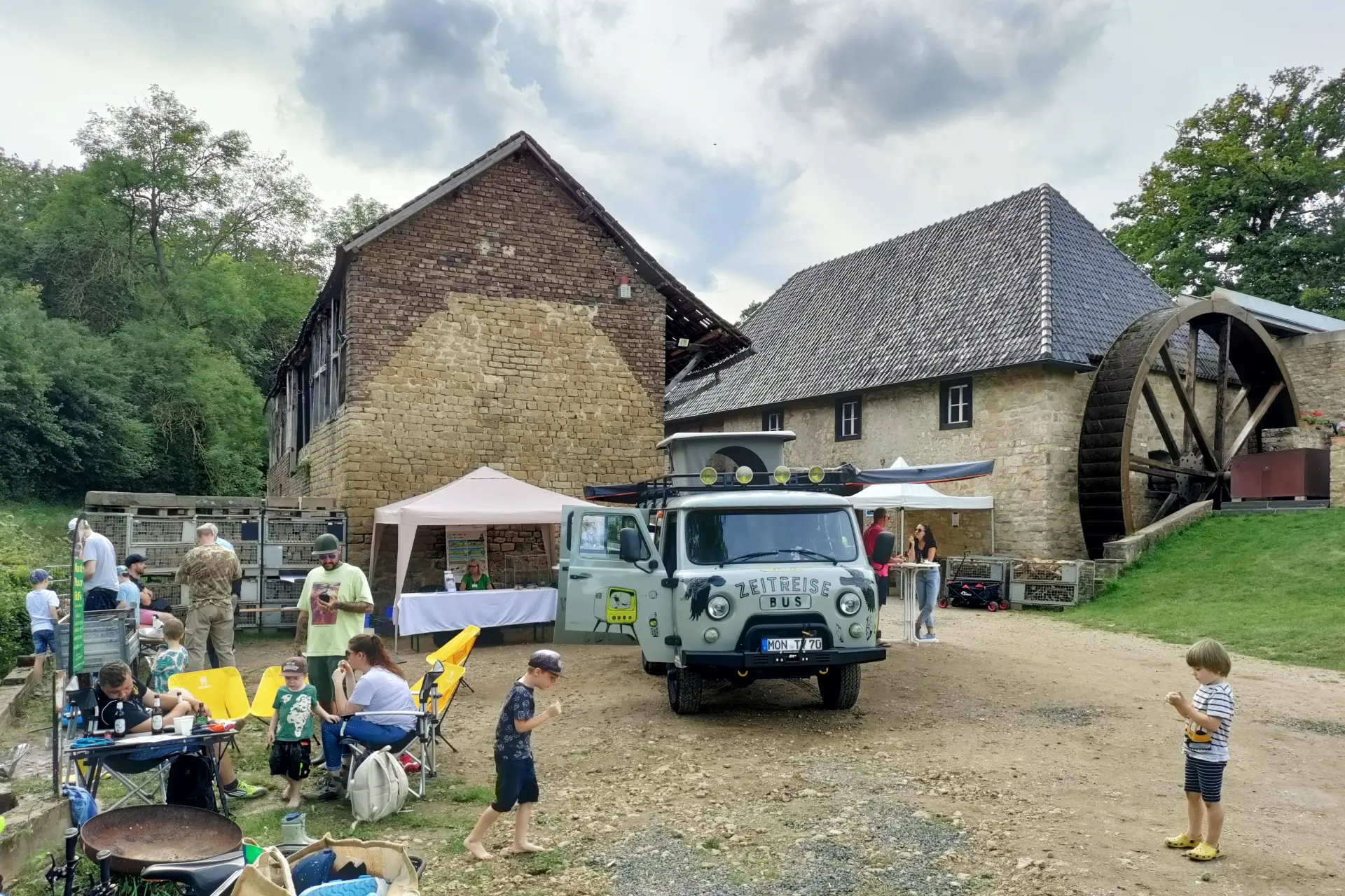 Das Foto zeigt im Hintergund zwei steinerne Gebäude. An einem gebäude ist ein großes Wasserrad zu sehen. Das Gebäude hat ein Schiefer-Dach. Im Bildzentrum ist ein Bus zu sehen, auf dem Zeitriese-Bus steht. Der Bus ist ein Oldtimer. Auf dem Foto sind mehrere personen zu sehen, die auf Camping-Stühlen sitzen oder über das Gelände gehen. 