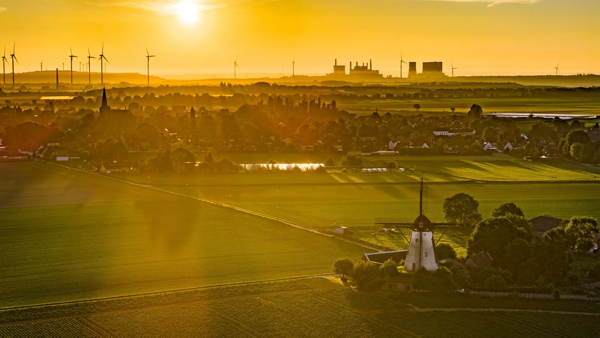 Das Bild zeigt einen Sonnenuntergang. Im Vordergrund ist eine WIndmühle zu sehen, Am Horizond sind Windenergieanlagen sowie Kraftwerke und Schornsteine zu sehen. 