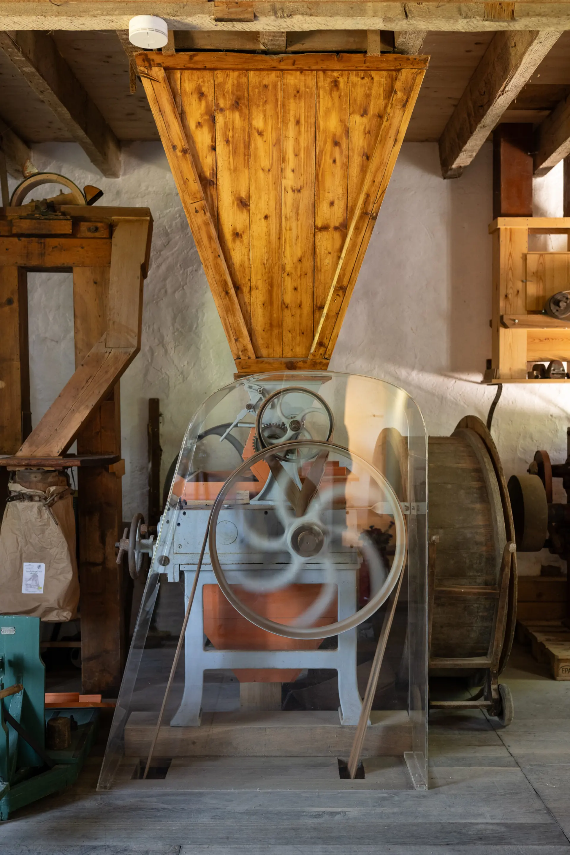 Das Foto zeigt einen hölzernen Trichter , der auf einer eisernen Maschine sitzt. An der Maschine ist ein Eisenrad mit einem Riemen zu sehen. Dieses Rad triebt die Maschine an. 