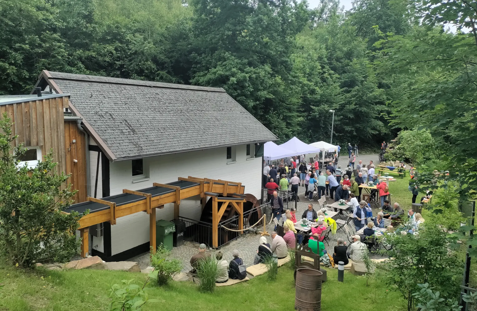 Rechts im Bild ist ein Mühlrad zu sehen, links ein Fachwerkhaus. Die Umgebung ist reich an Bäumen und Vegetation. 