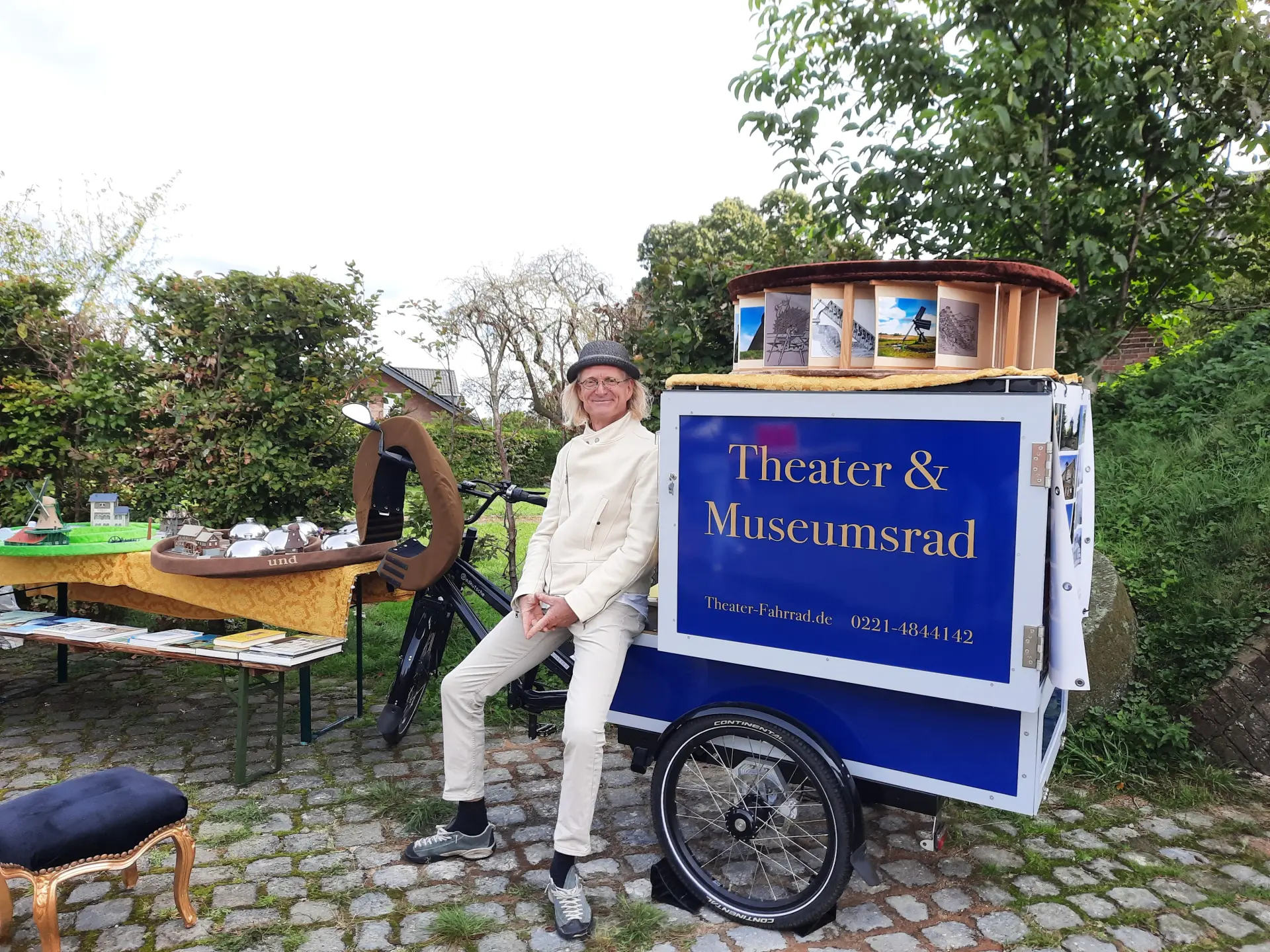 Das Foto zeigt einen Mann in weißem Anzug, der an das "Theater & Museumsrad" lehnt. Auf diesem Fahrrad liegt ein Modell eines Mühlrades. 