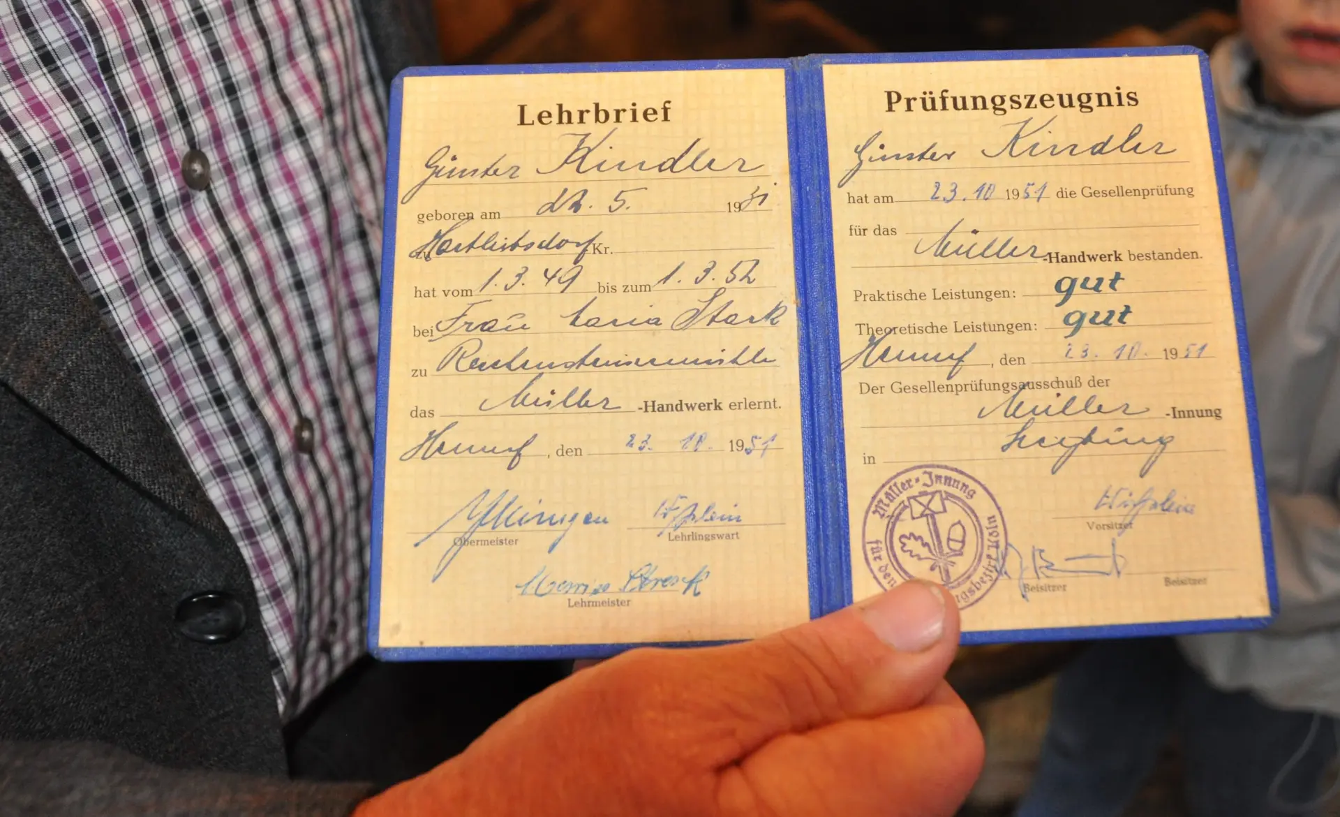 Das Foto zeigt einen "Lehrbrief" zum Müller. Es ist ein zweiseitiges Dokument auf gelbem Papier. Darauf stehen handschriftliche EInträge und es gibt einen Stempel. Der Lehrbrief wird von der Hand eines älteren Mannes gehalten. 