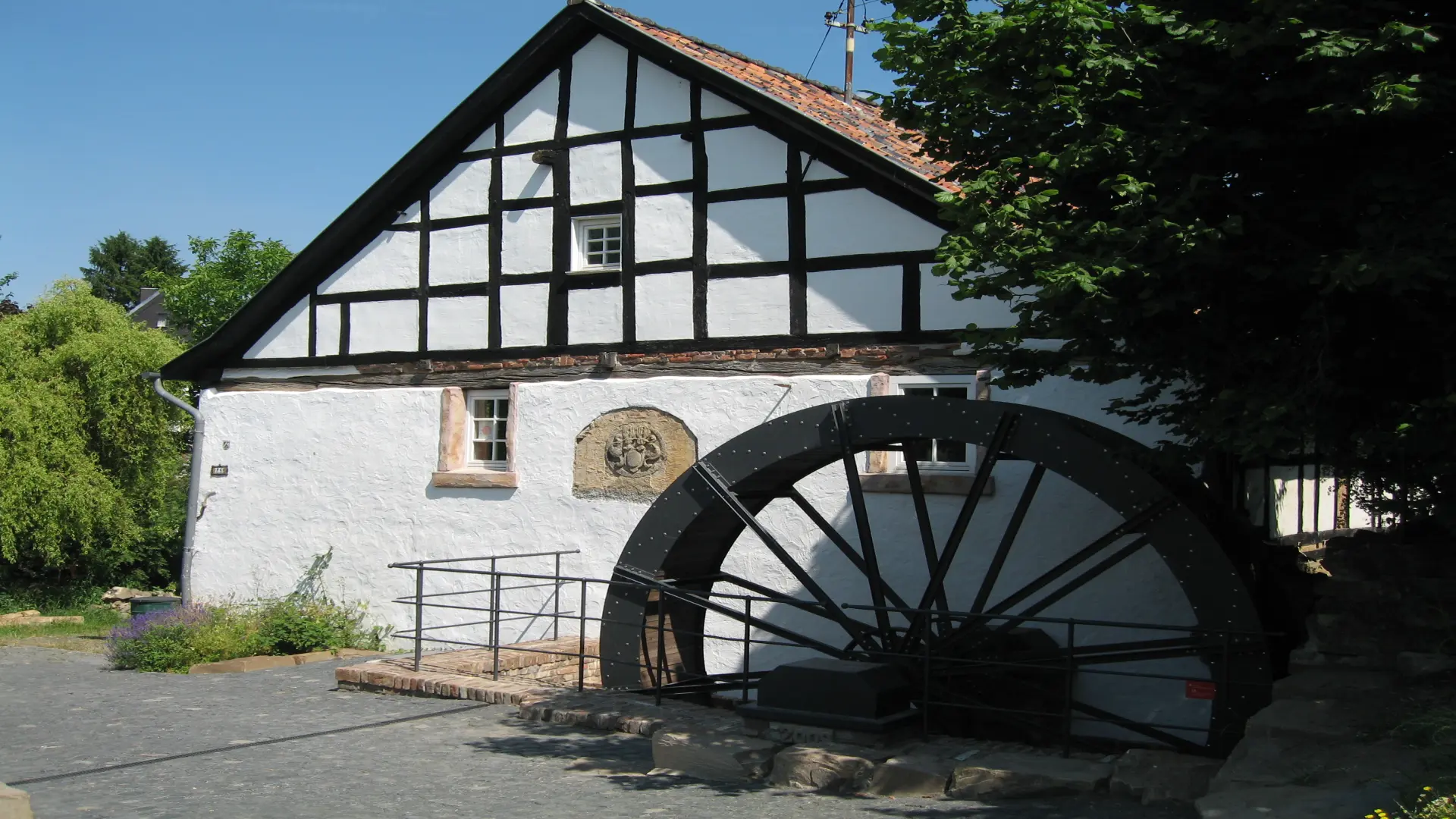 Foto: Fachwerkgebäude mit Mühlrad aus Eisen. Im Vordergrund Kopfsteinpflaster.