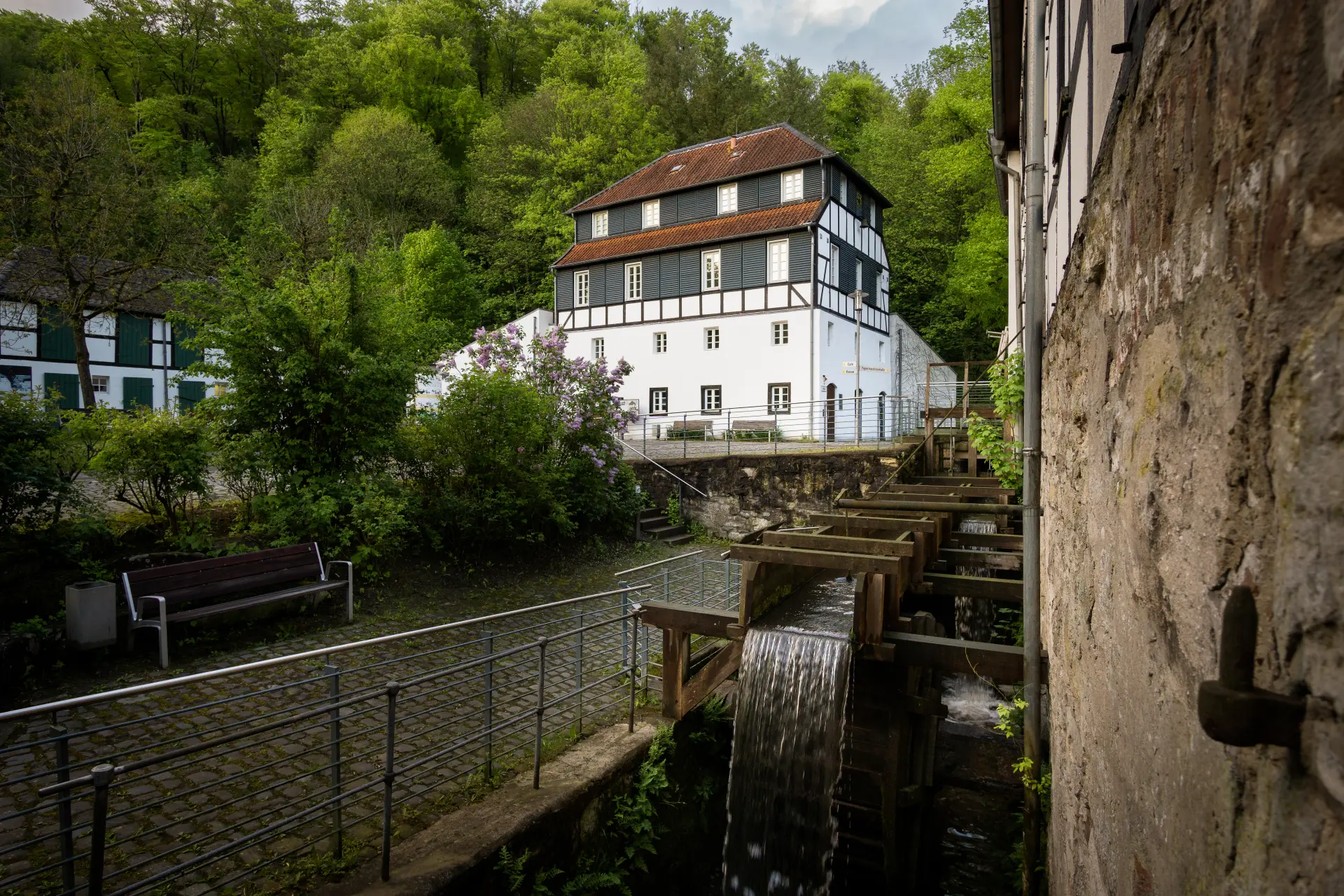 Foto: Mehrstöckiges Gebäude mit weißer Fassade. Die Obergeschosse sind aus Fachwerk. Im Vordergrund ist ein Mühlrad zu sehen.