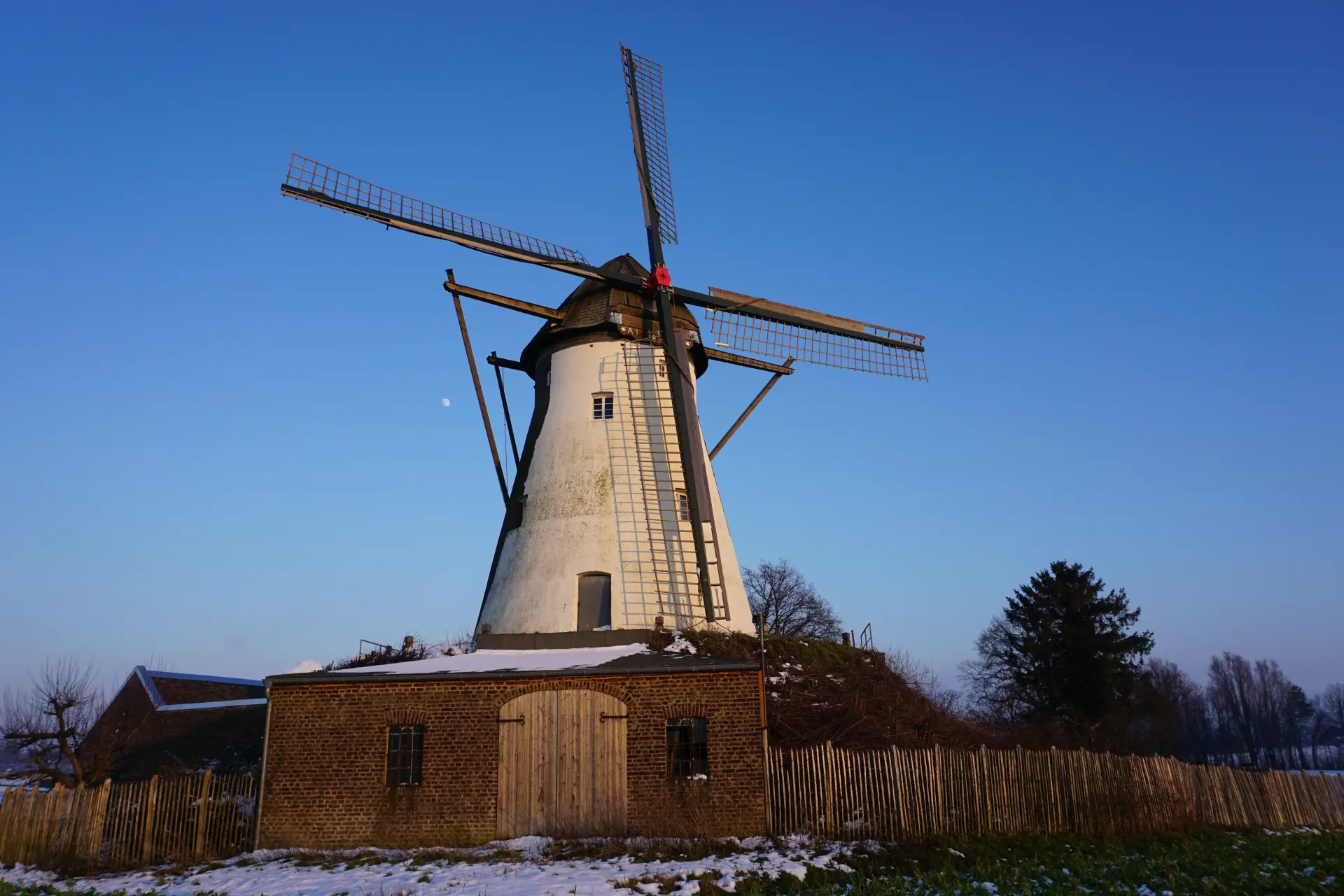 Windmühle mit vier Flügeln im Abendlicht. Im Vordergrund sieht man etwas Schnee auf dem Boden liegen, der Mond steht am Himmel. 
