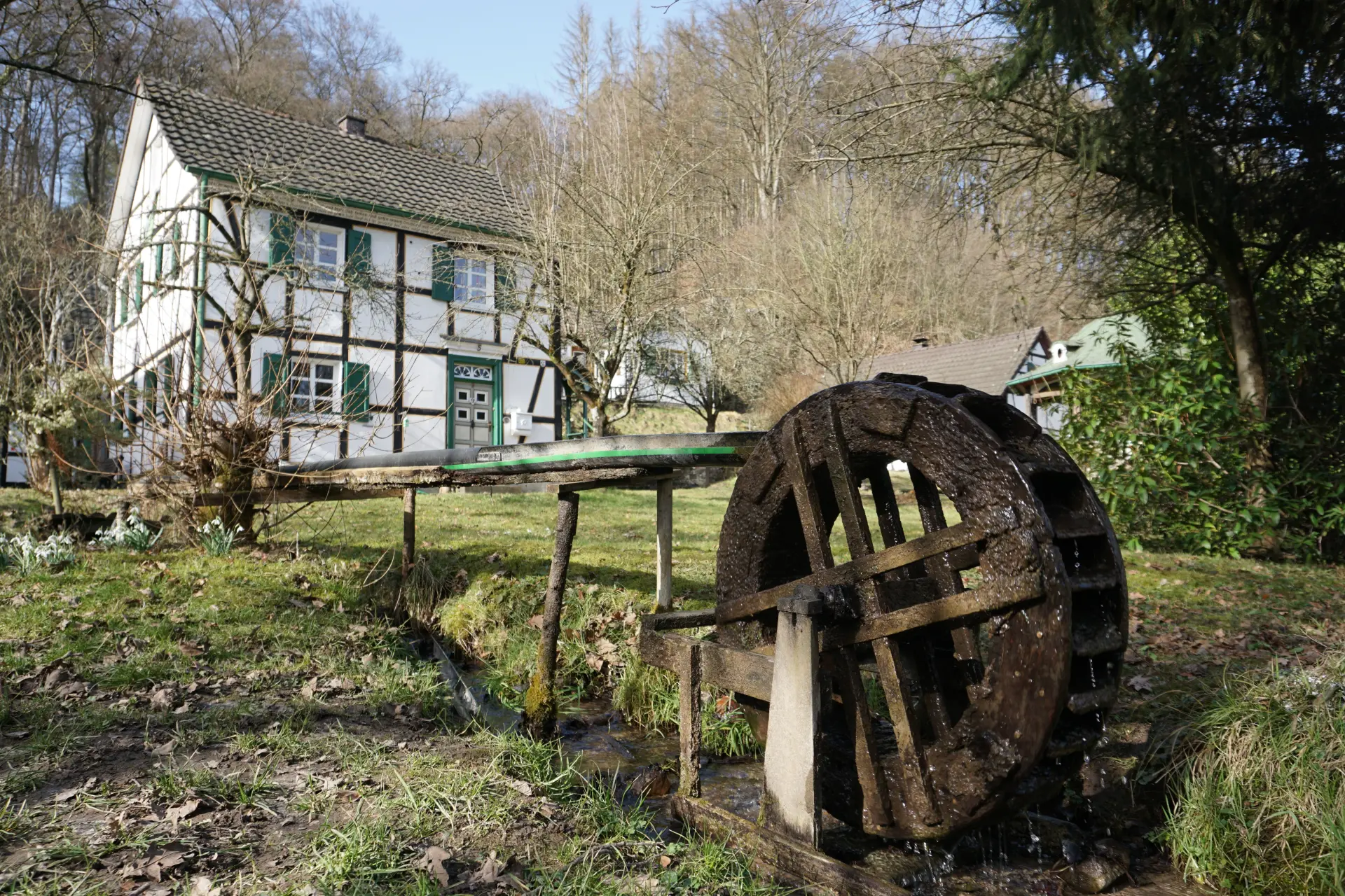 Rechts im Bild ist ein Mühlrad zu sehen, links ein Fachwerkhaus. Die Umgebung ist reich an Bäumen und Vegetation. 