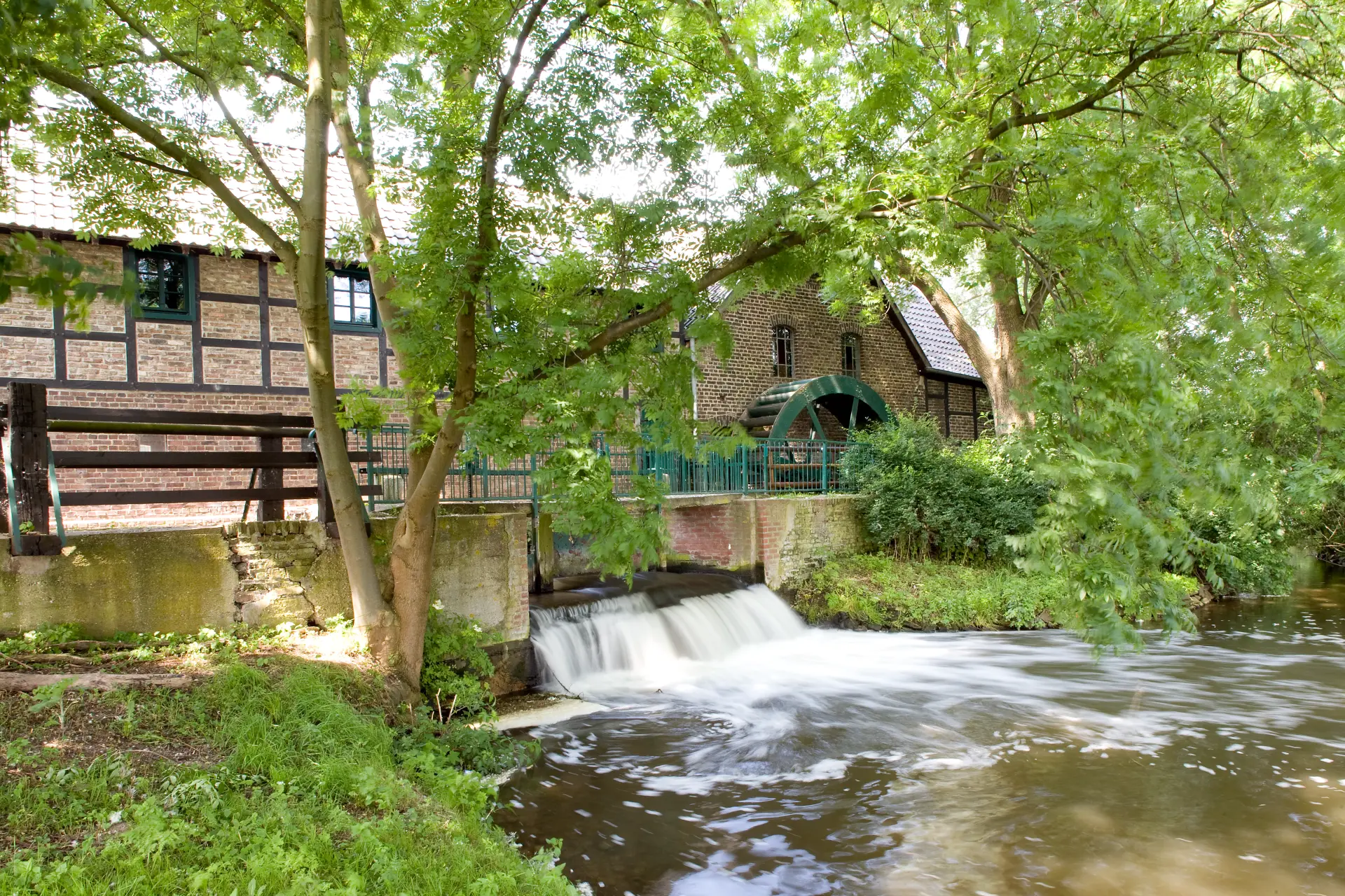 Das Foto zeigt eine Mühle mit Mühlrad. Sie steht an einem Fluss. Im Umfeld sind Bäume und Büsche zu sehen. 