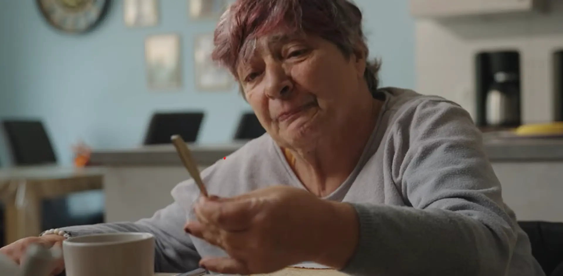 Szene aus dem Film: Eine Frau sitzt am Tisch mit einer Kaffeetasse vor sich und schaut auf den Löffel in ihrer Hand