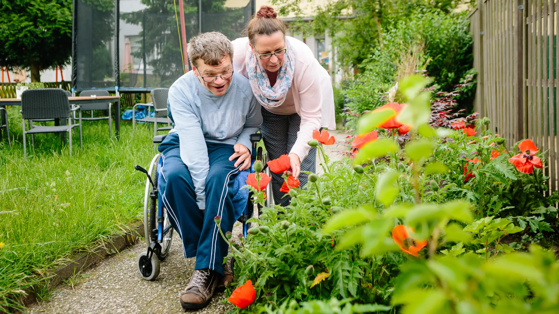 Auf dem Bild zu sehen sind ein Mann im Rollstuhl mit seiner Betreuerin vor einem Blumenbeet