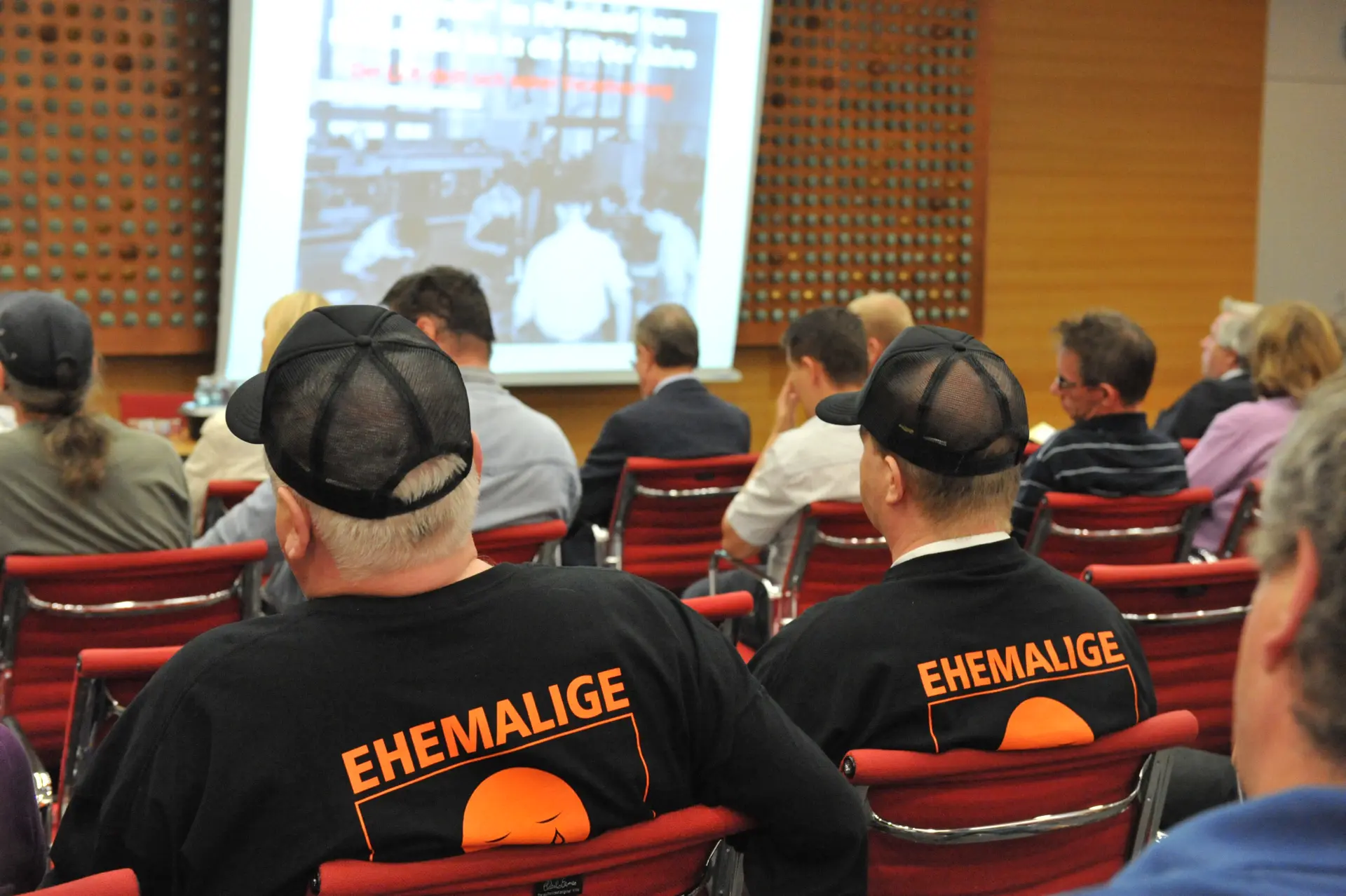 Viele Personen gucken auf eine Leinwand vor ihnen. Man sieht die Personen von hinten. Auf ihren T-Shirts steht in orangener Schrift "Heimkinder".