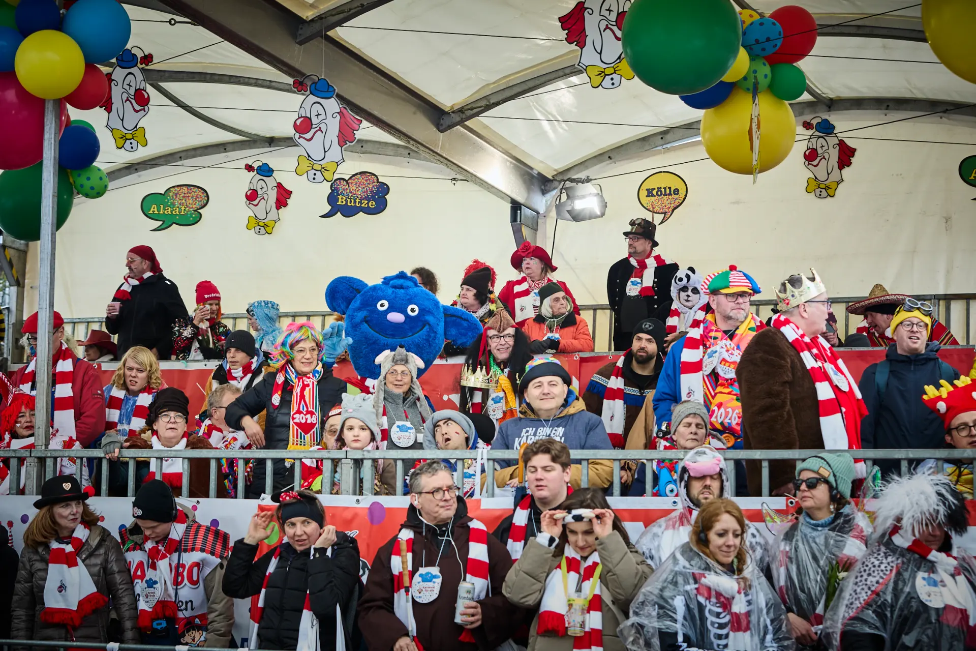 Viele Menschen und das LVR-Maskottchen Mitmän, ein blauer Kobold, feiern zusammen auf der Tribüne beim Kölner Rosenmontagszug.