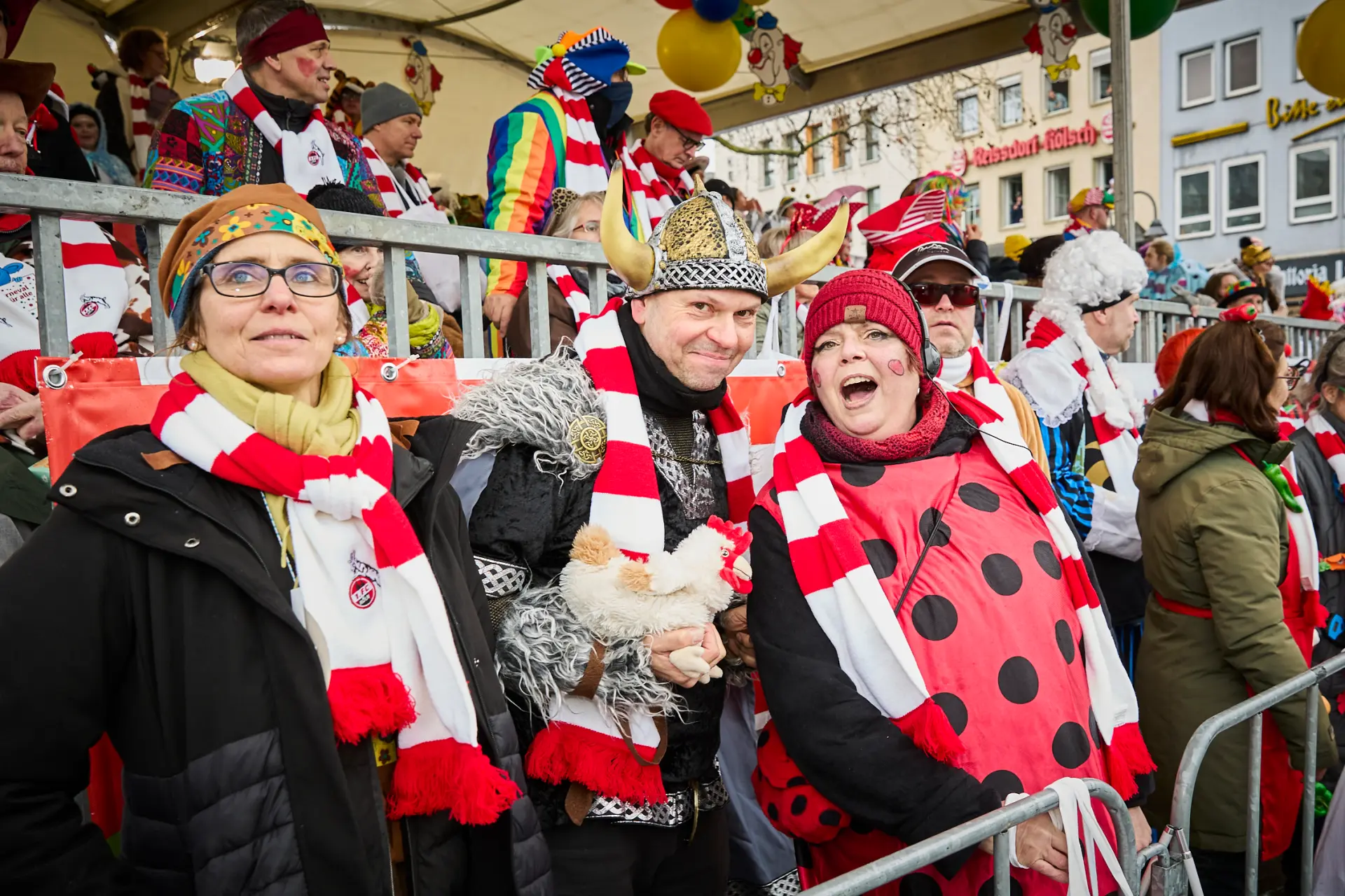 Drei feiernde Menschen tragen rot-weiß-getreifte Schals des 1. FC Kölns und schunkeln gemeinsam auf der Tribüne beim Rosenmontagszug in Köln.