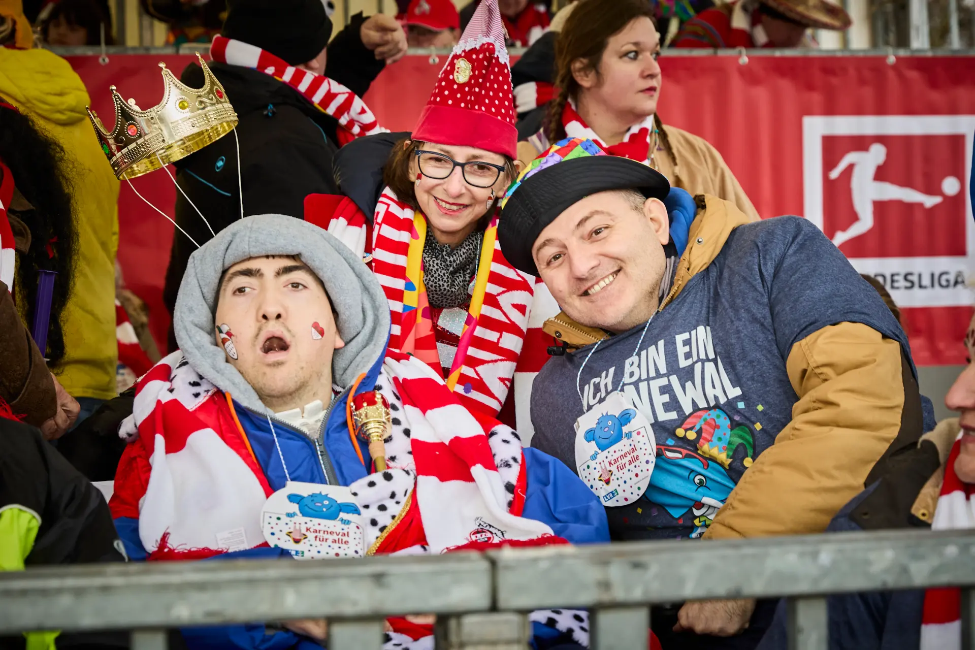 Zwei Männer mit und ohne Behinderung tragen rot-weiß-gestreifte Schals des 1. FC Kölns und feiern beim Rosenmontag auf der Tribüne.