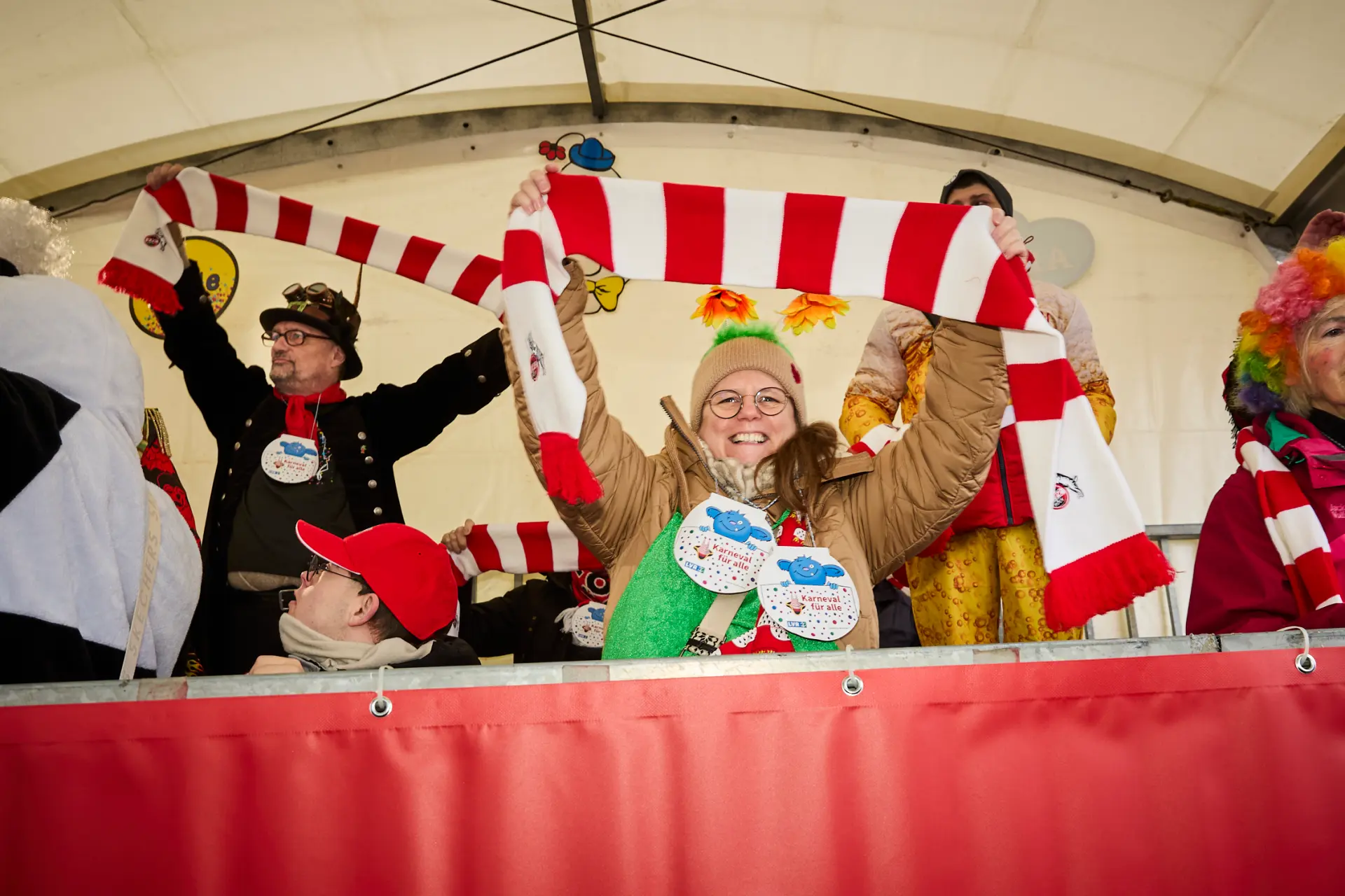 Eine gutgelaunte Frau sitzt auf der Tribüne beim Kölner Rosenmontagszug und hält einen rot-weiß-gestreiften Schal des 1. FC Köln in die Höhe.
