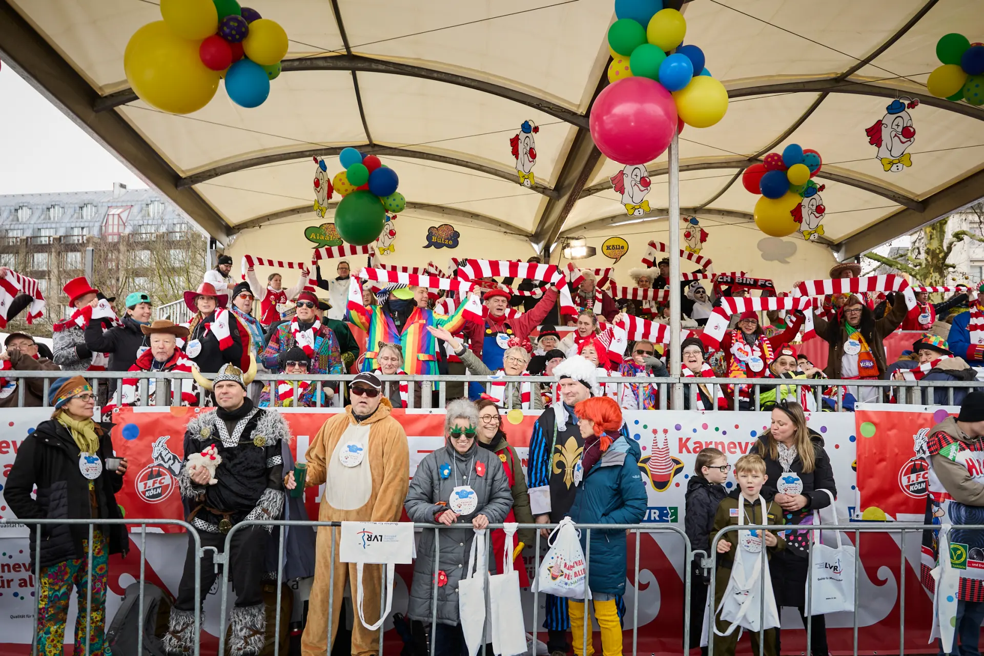 Die Tribüne beim Kölner Rosenmontagszug ist voller feiernder Menschen in Kostümen und in rot-weiß-gestreiften Schals des 1. FC Köln.