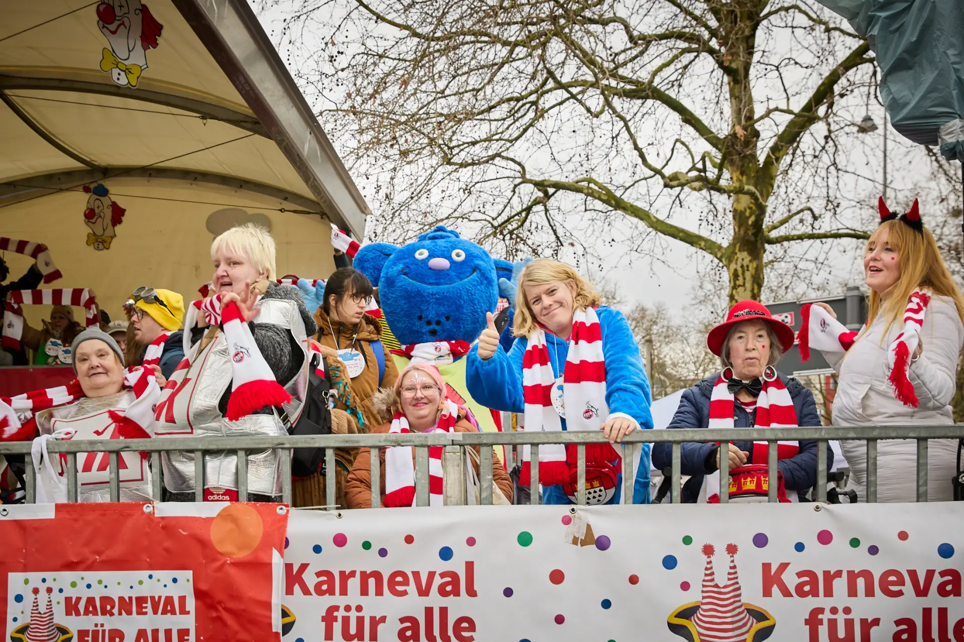 Gut gelaunte Menschen stehen zusammen mit dem LVR-Maskottchen Mitmän, einem blauen Kobold, auf der Tribüne und feiern.