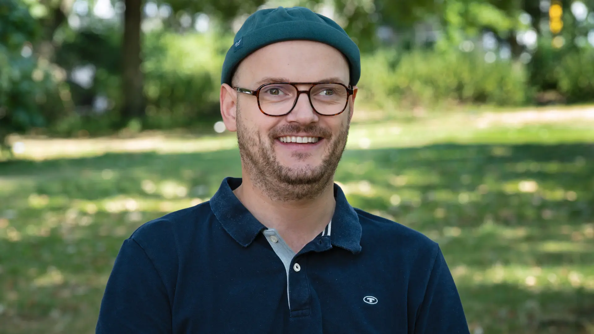 Porträt eines lächelnden Mannes mit Brille, kurzem Bart, petrolfarbener Mütze und dunkelblauem Poloshirt. Im unscharfen Hintergrund sind eine Wiese und Bäume zu sehen.