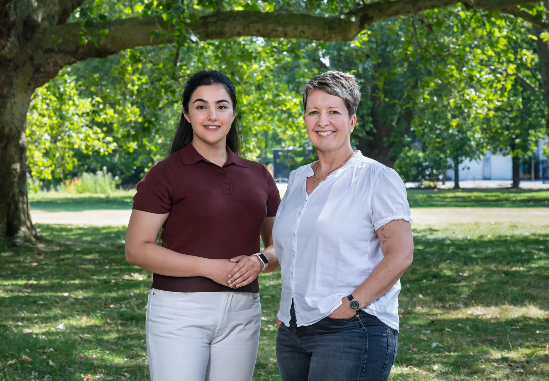 Zwei Frauen stehen nebeneinander unter einem großen Baum und lächeln in die Kamera. Die Frau links hat lange dunkle Haare und trägt ein bordeauxrotes Poloshirt und eine helle Hose. Die Frau rechts hat kurze braun-graue Haare und trägt eine weiße Bluse und dunkle Jeans.