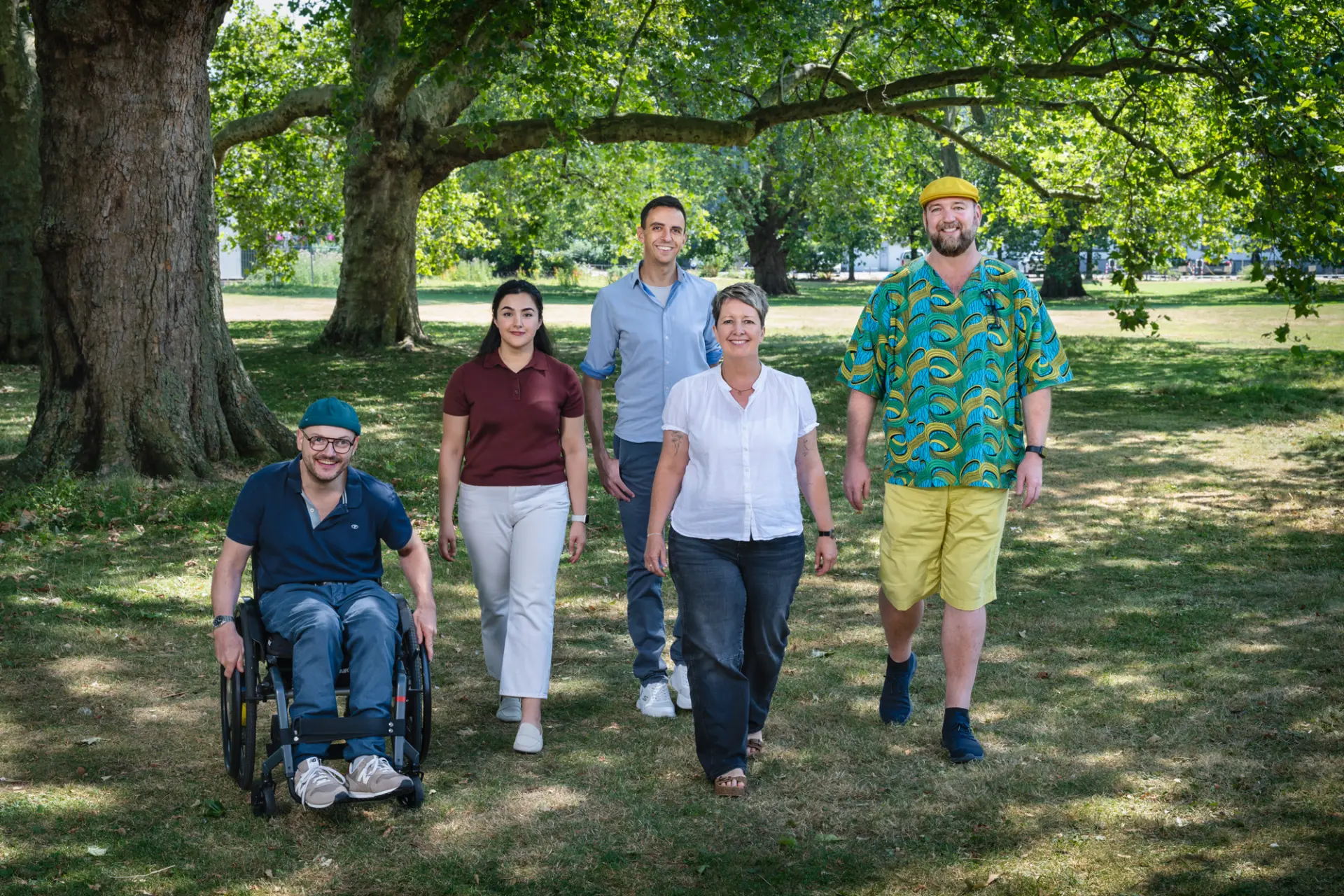 Fünf Personen gehen gemeinsam durch einen grünen Park unter großen Bäumen, eine Person nutzt einen Rollstuhl.
