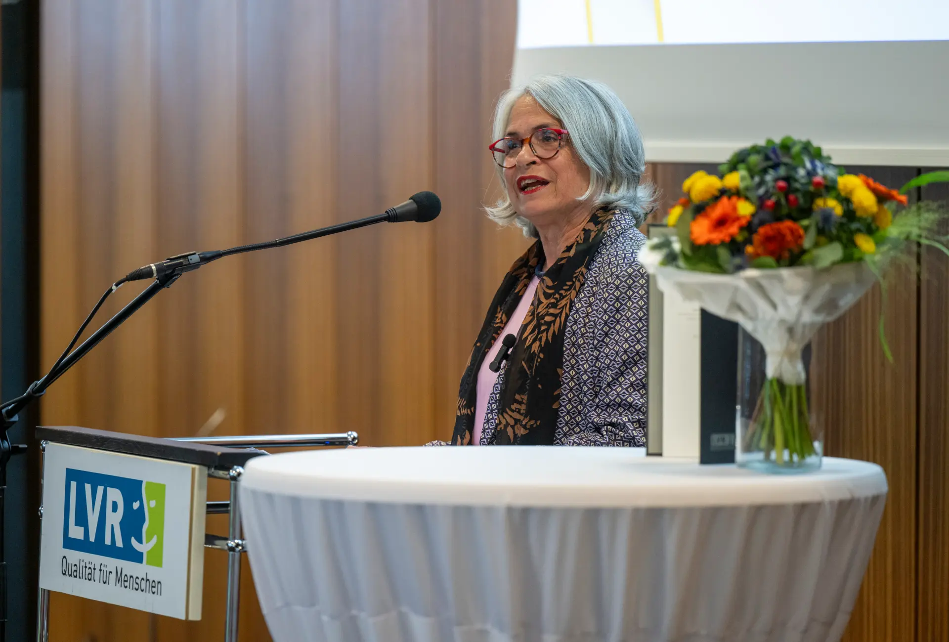 Eine Frau mit grauen schulterlangen Haaren und roter Brille steht an einem Rednerpult.
