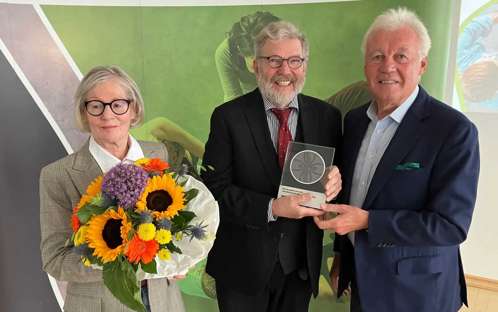 Eine Frau hält einen Blumenstrauß. Daneben rechts stehen zwei Männer, die eine Trophäe in der Hand halten.