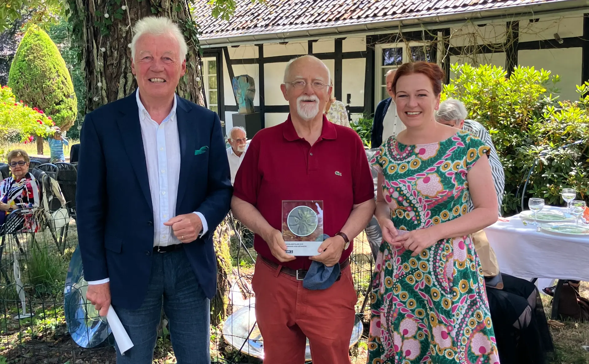 Zwei Männer und eine Frau stehen im Garten. Der Mann in der Mitte hält eine Trophäe in der Hand.