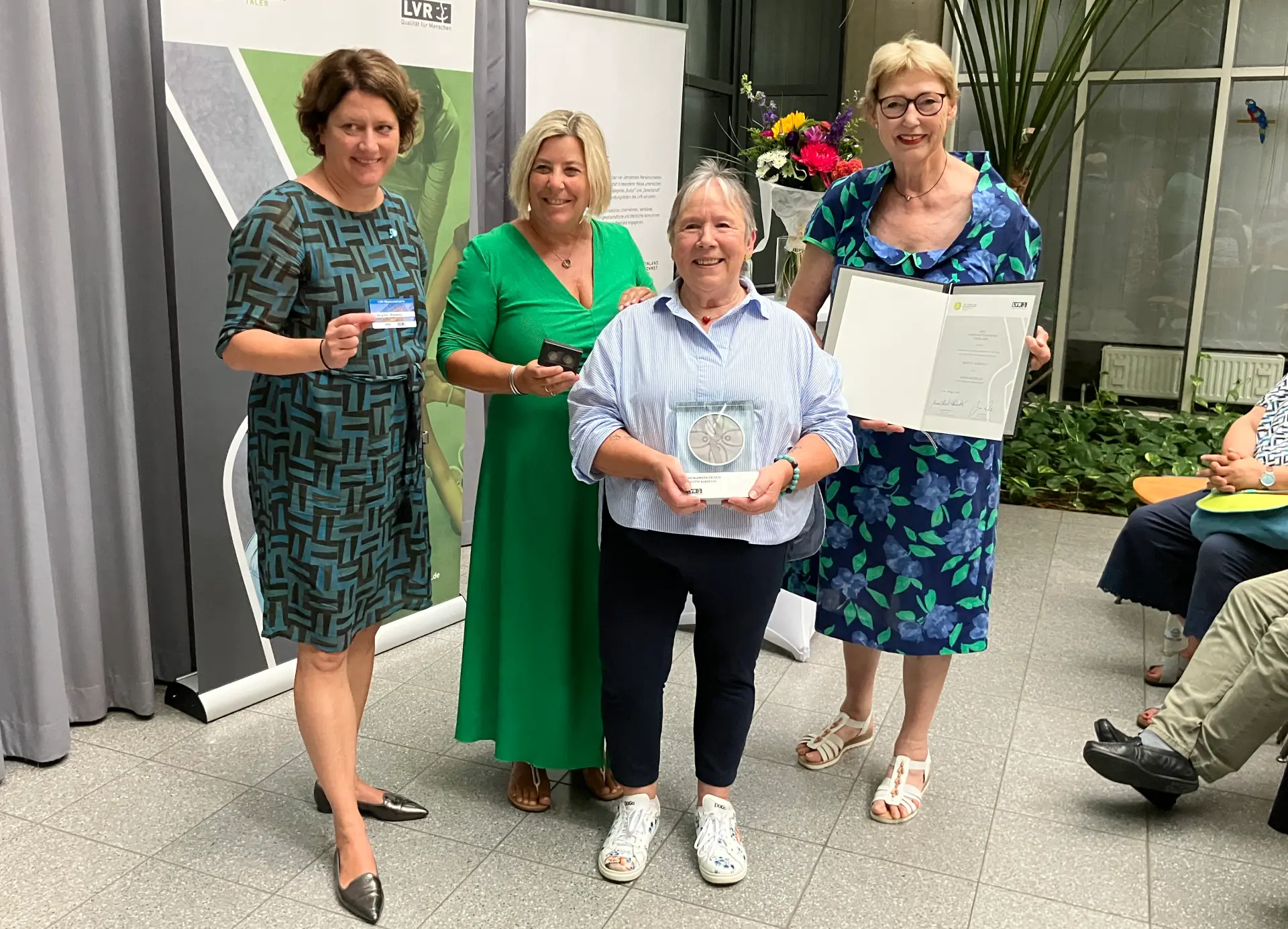 Vier Frauen lächeln in die Kamera. Die Frau ganz rechts hält eine Urkunde hoch. Die Frau links daneben hält eine Trophäe in der Hand.