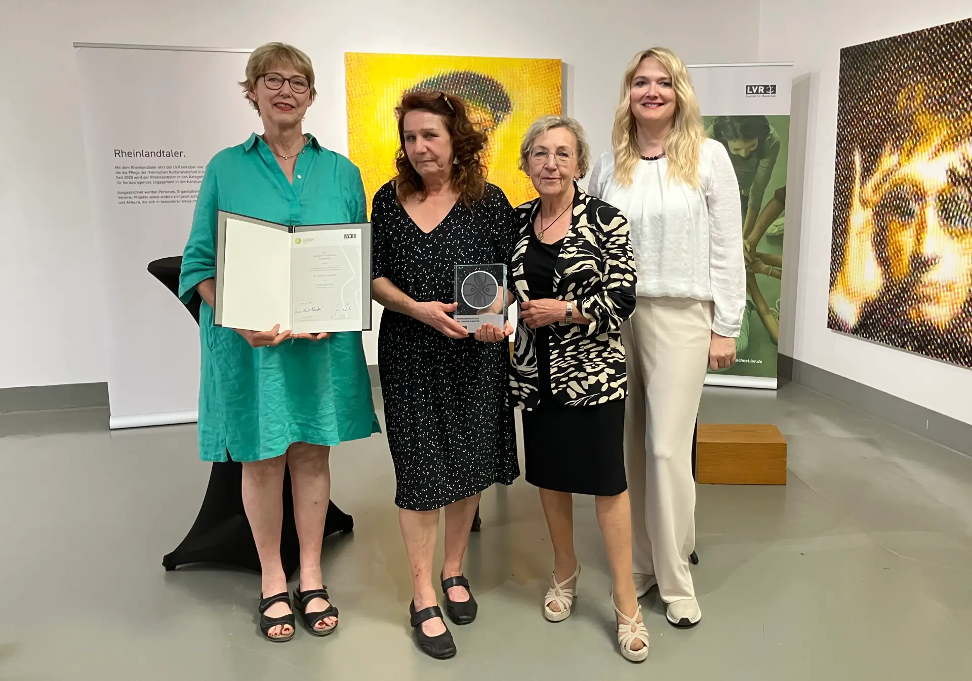 Vier Frauen. Die Frau ganz links hält eine Urkunde. Die Frau rechts daneben hält eine Trophäe.