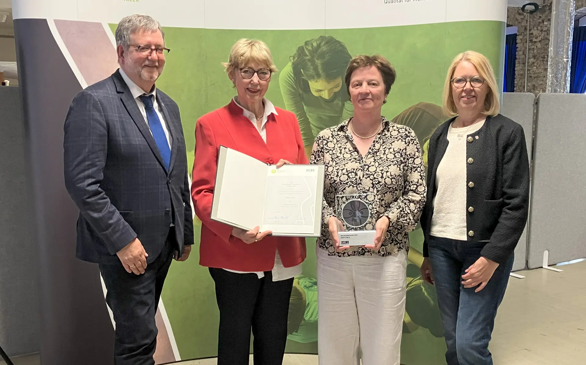Ein Mann und drei Frauen. Eine Frau hält eine Urkunde in der Hand, eine weitere hält eine Trophäe.