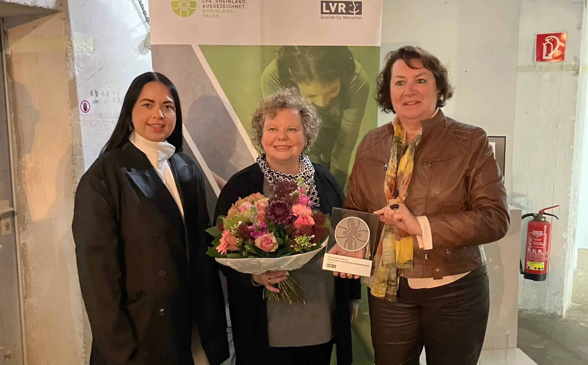 Drei Frauen stehen vor einer Leinwand mit dem Rheinland ausgezeichnet-Logo. Die Frau in der Mitte hält einen Blumenstrauß in der Hand. Die Frau links daneben eine Trophäe.