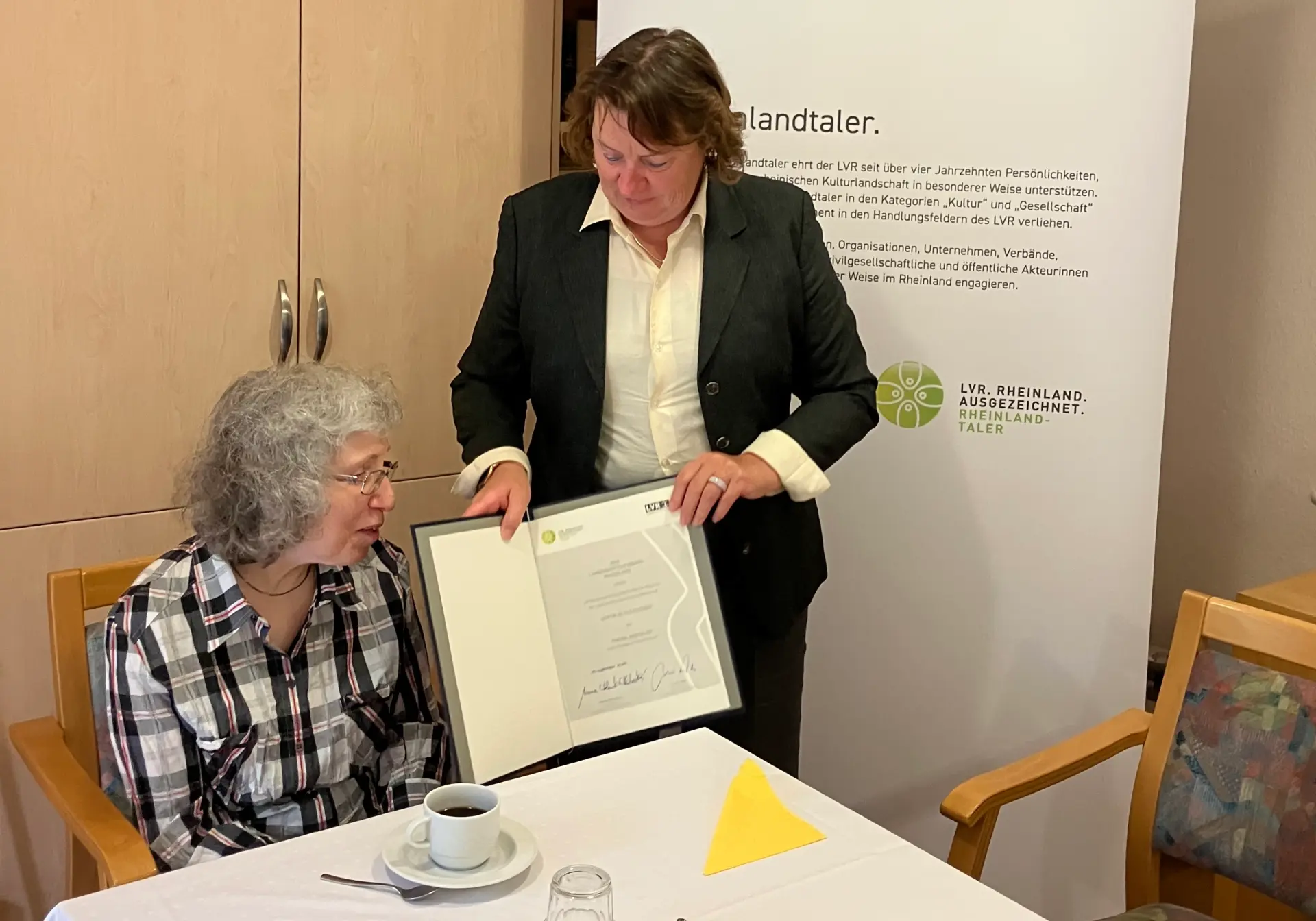 Eine Frau sitzt am Tisch. Daneben steht eine Frau, die ihr eine Urkunde überreicht.
